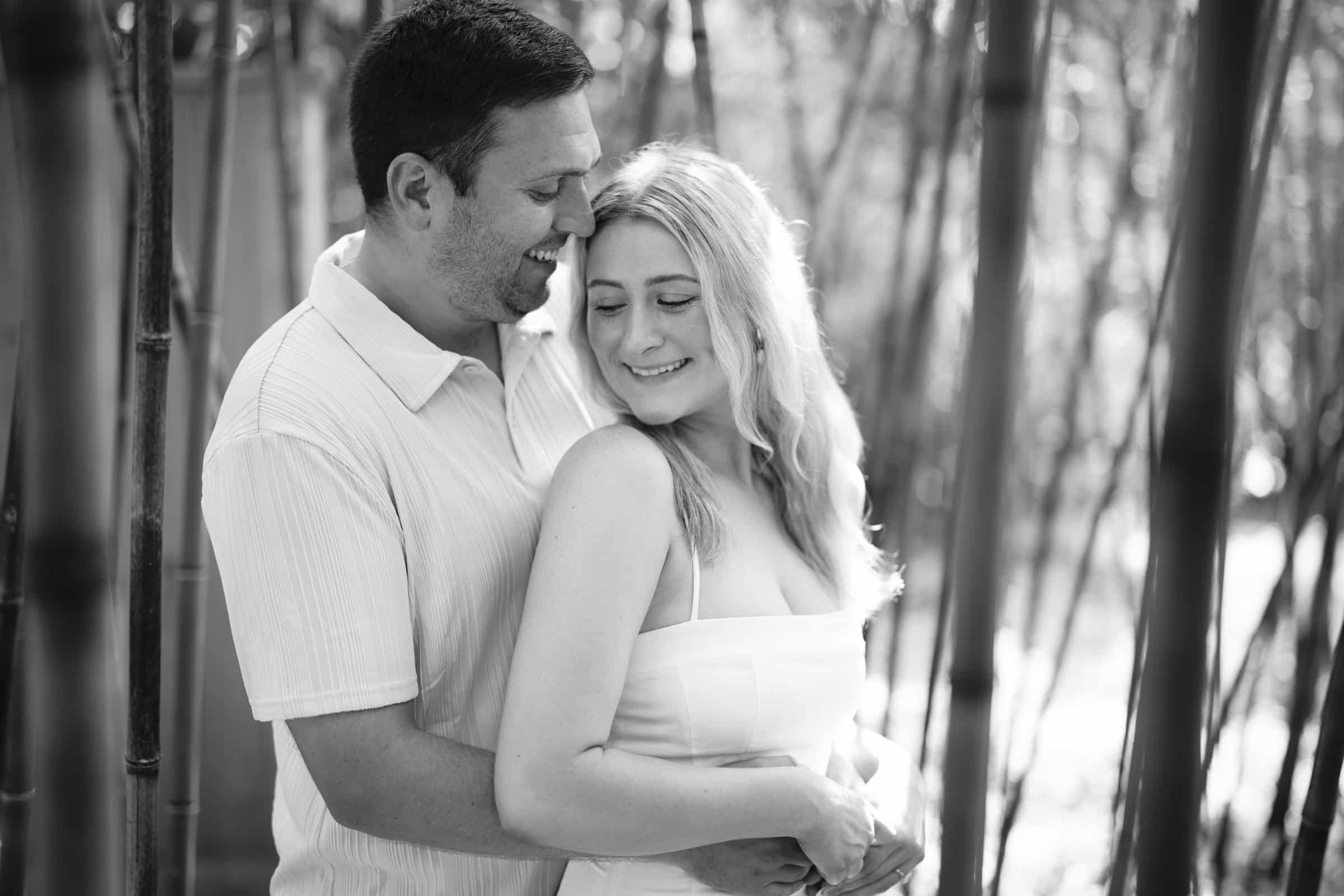 A man and woman stand close together smiling among tall bamboo stalks. The woman faces forward with her eyes closed, while the man stands behind her, embracing her and looking at her. The image is in black and white.