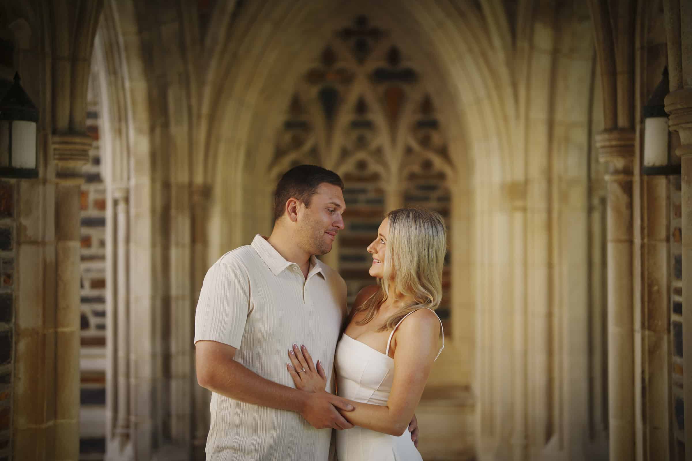 A couple stands closely together, gazing at each other and smiling, in an ornate stone archway with soft, warm lighting. The woman wears a white dress and the man wears a light-colored shirt.