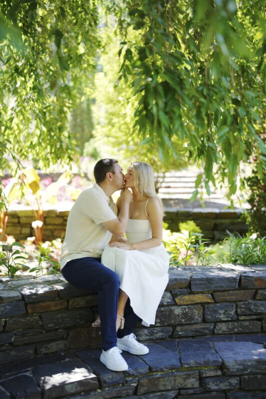 A couple sits on a stone wall in a lush garden, sharing a tender kiss. The woman wears a white dress and sneakers, while the man wears a light shirt and dark pants. Green leaves hang above them, creating a serene, romantic scene.