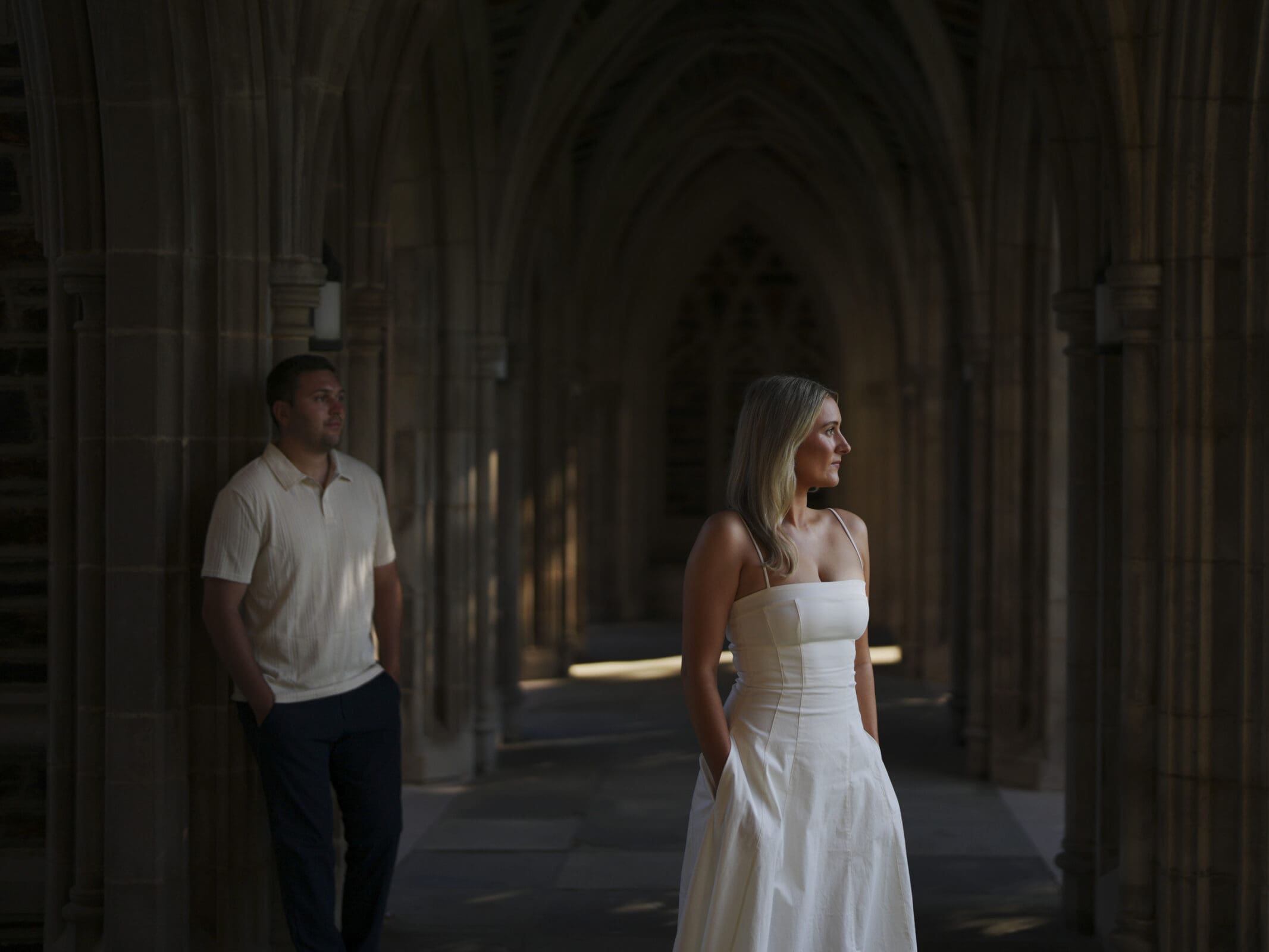 A woman in a white dress stands in a stone hallway with arched ceilings, looking to the side. A man in a white shirt and dark pants stands in the background, partially in shadow, with hands in his pockets.