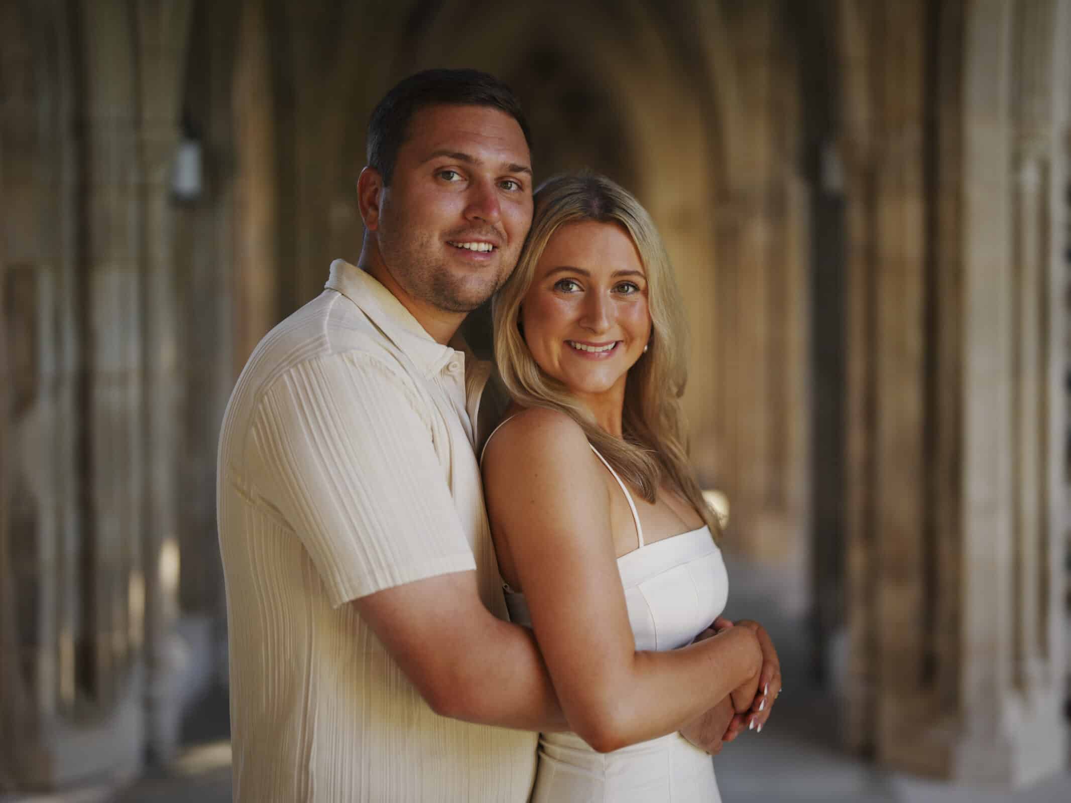 A smiling couple embraces in a sunlit stone corridor with arched ceilings. The man wears a light short-sleeve shirt and the woman wears a white dress. Both look at the camera, standing close together.