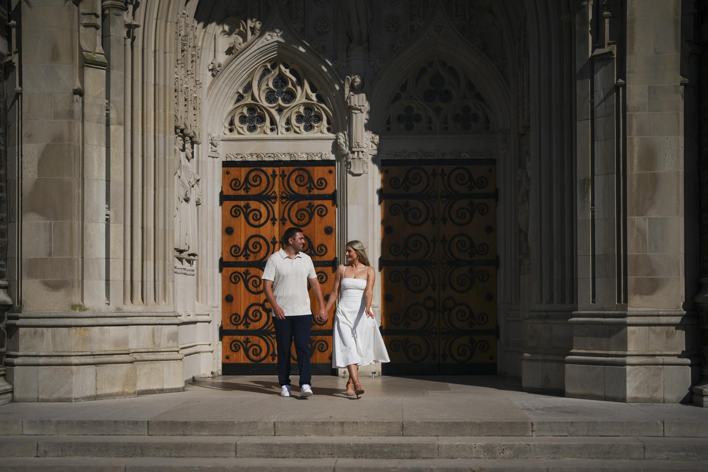 A man and woman hold hands while standing in front of ornate wooden doors with black iron designs, set in a large stone building with Gothic architectural details.
