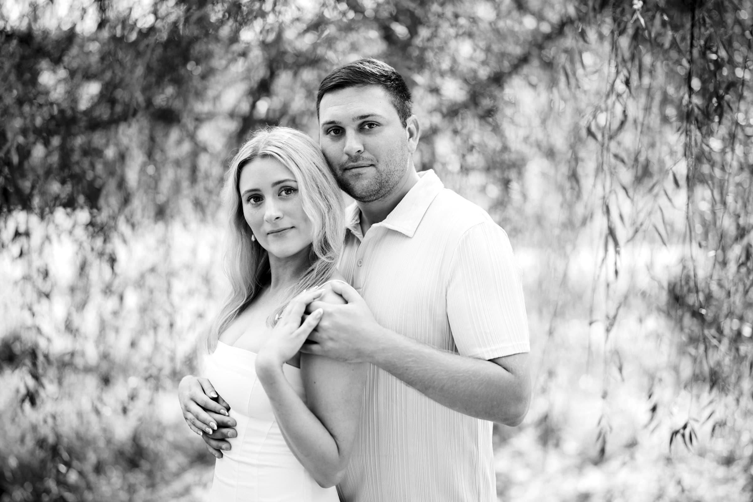 A couple stands close together outdoors, holding hands and looking at the camera, surrounded by hanging tree branches and soft natural light in a serene setting.