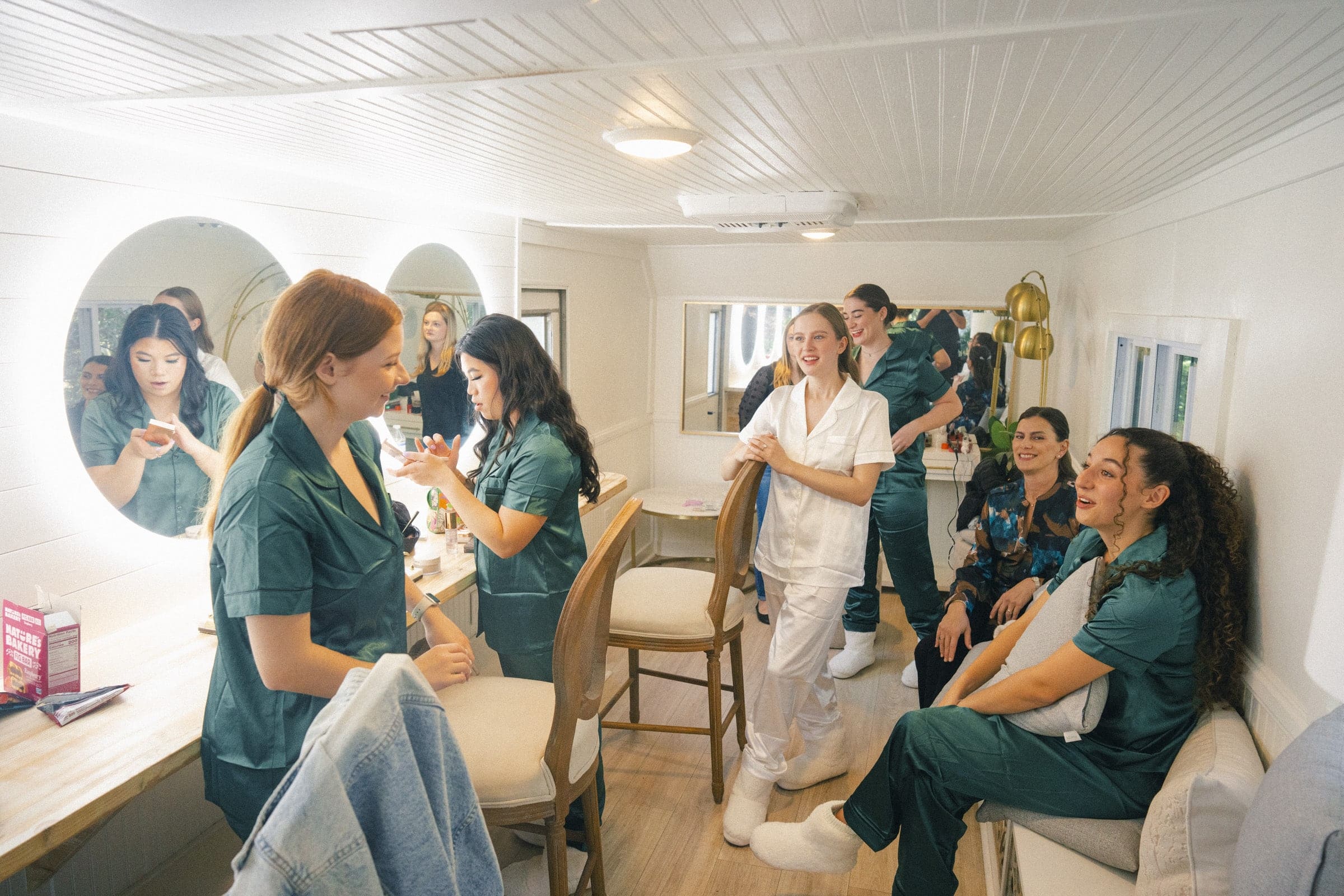 A group of women in green and white uniforms laugh and chat in a well-lit dressing room with mirrors, stools, and makeup on the counter. Some are seated while others stand, creating a lively and friendly atmosphere.