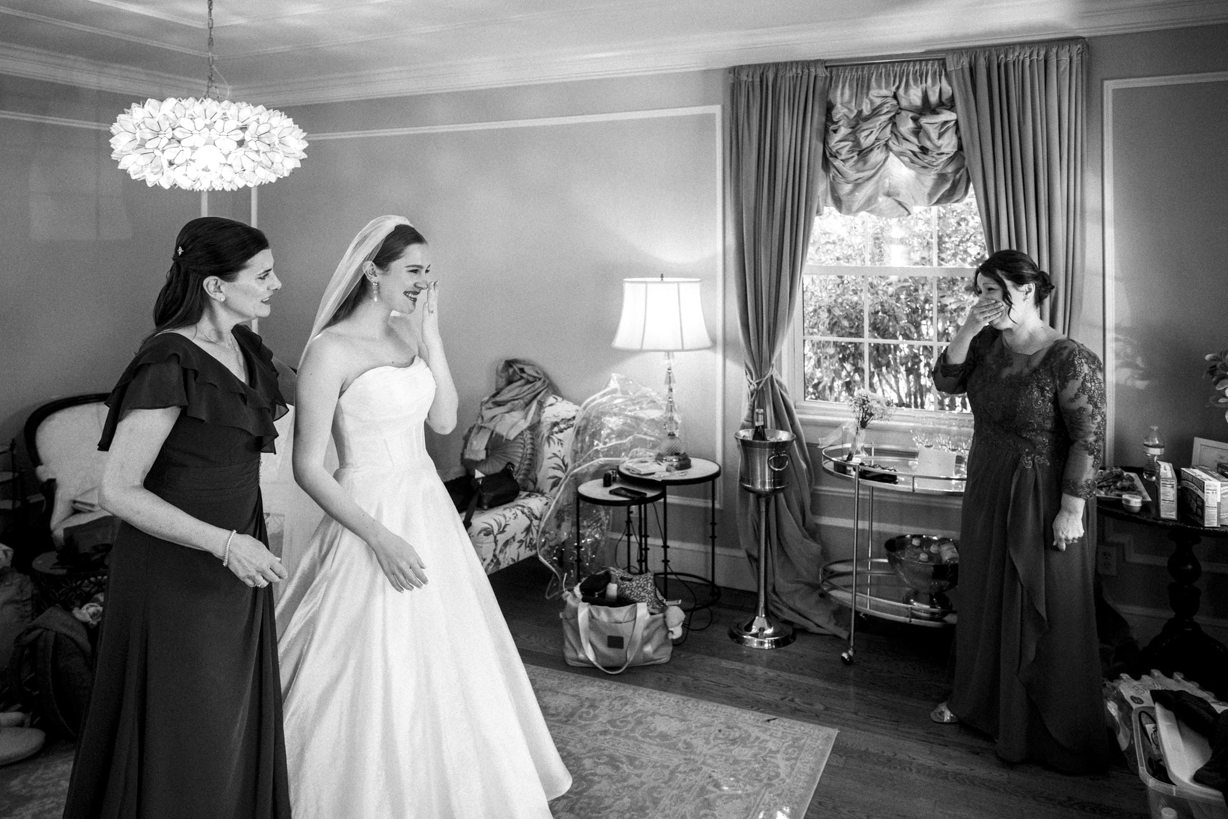 A bride in a wedding dress smiles and wipes away a tear, standing with two women in formal gowns in a bright, elegant room with a large window and ornate decor.