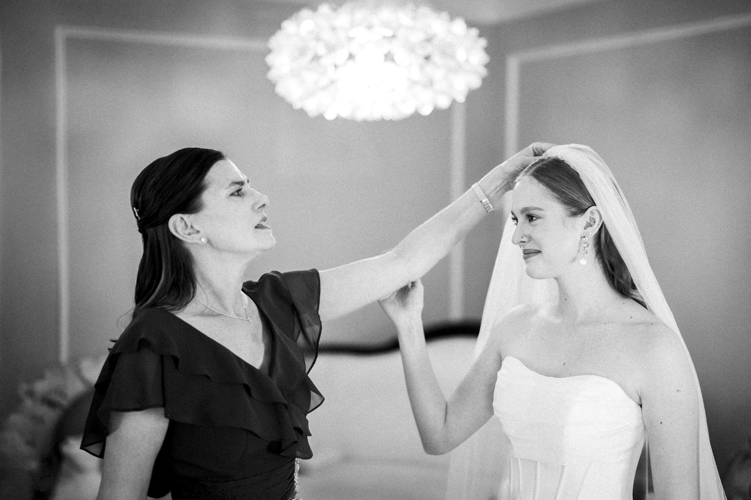 A woman adjusts a brides veil as they stand together indoors. The bride wears a strapless wedding dress and smiles softly, while the woman appears focused on fixing the veil. The room is softly lit with a chandelier above.