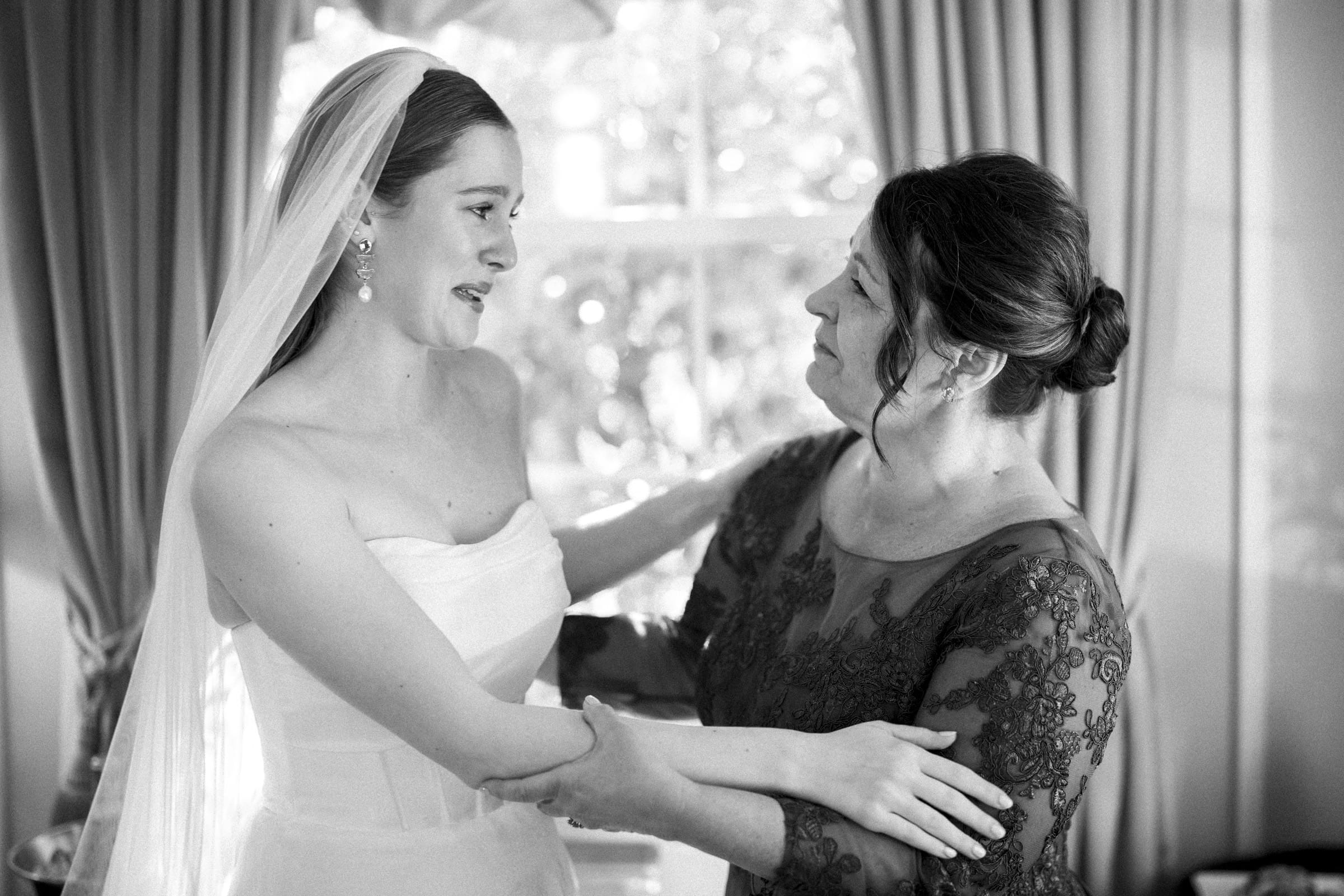 A bride in a strapless dress and veil smiles warmly as she holds hands with a woman in an elegant lace dress. They share a tender moment indoors, framed by soft light and curtains.