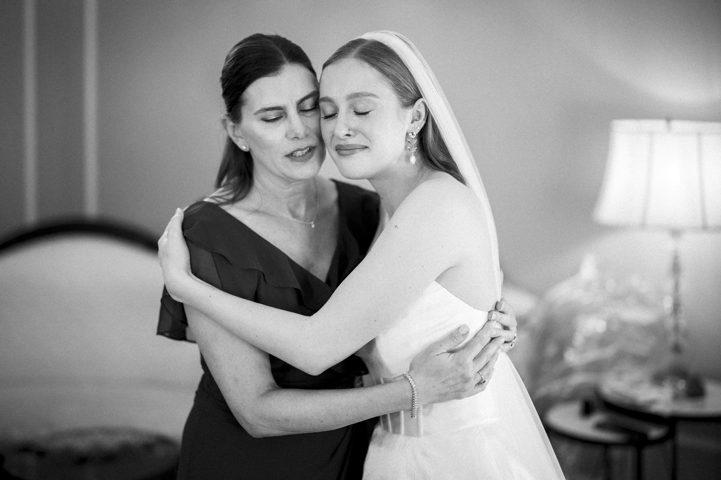 A bride in a wedding dress and veil hugs a woman in a dark dress, both with eyes closed, sharing an emotional moment in a softly lit room with elegant furniture in the background.