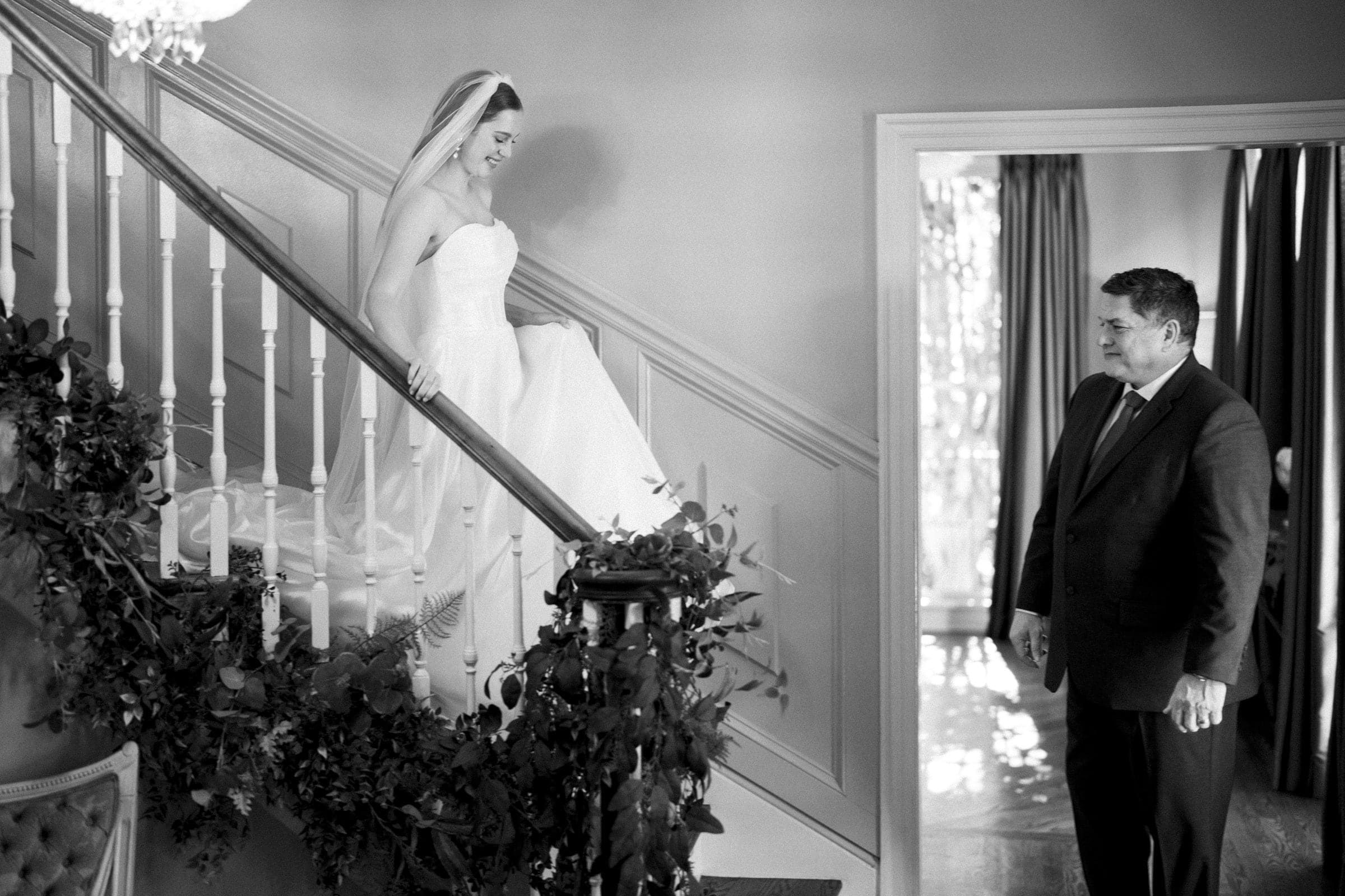 A bride in a wedding gown descends a staircase decorated with greenery, smiling at a man in a suit who stands at the bottom, watching her with a proud expression in a warmly lit room.