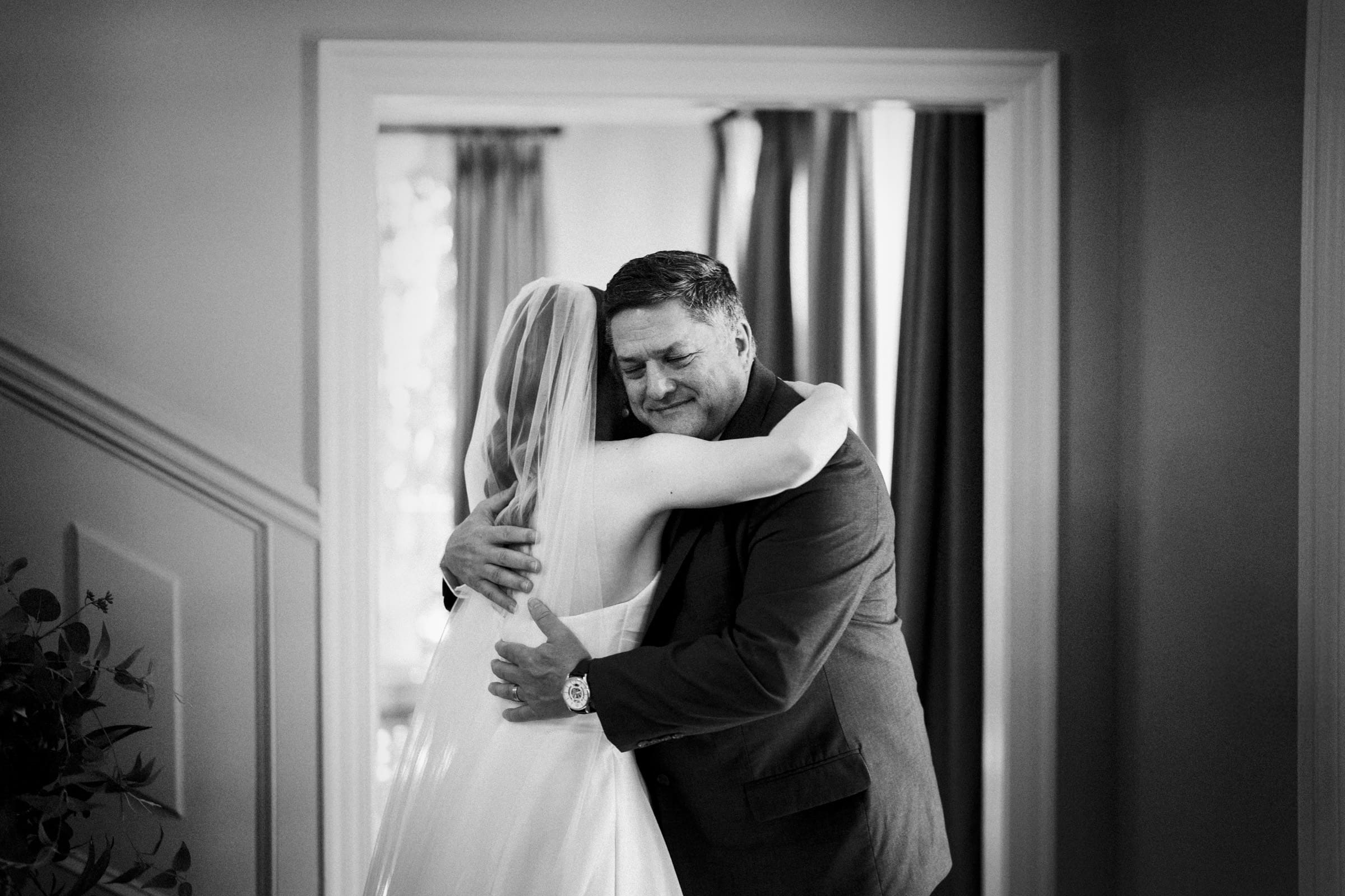 A bride in a wedding dress and veil hugs an older man in a suit, possibly her father, in a warmly lit room. Both appear emotional and happy, sharing a heartfelt moment.