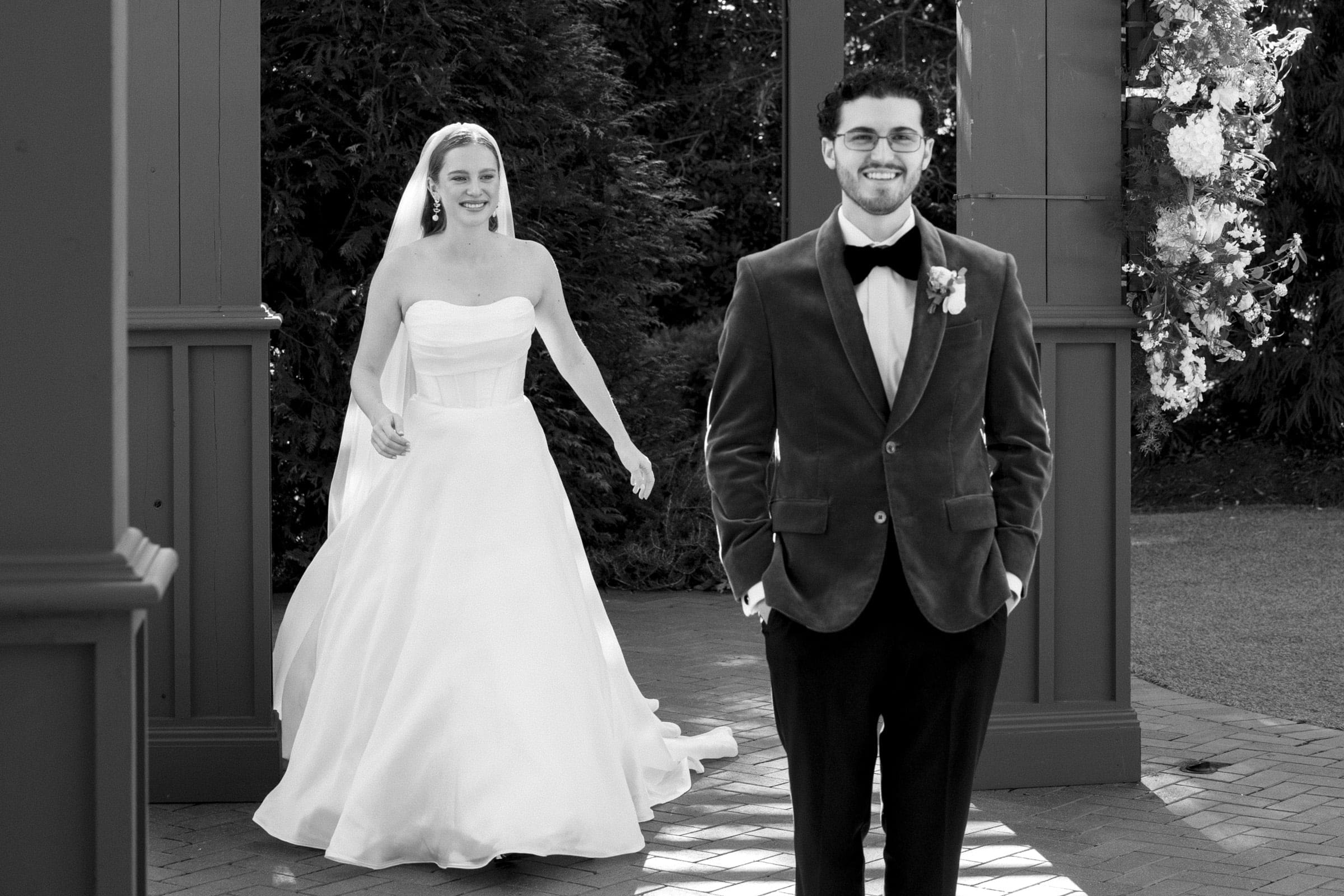 A bride in a strapless gown smiles as she walks toward a groom in a suit jacket with his hands in his pockets, during their outdoor wedding. Both are happy and the scene is in black and white.
