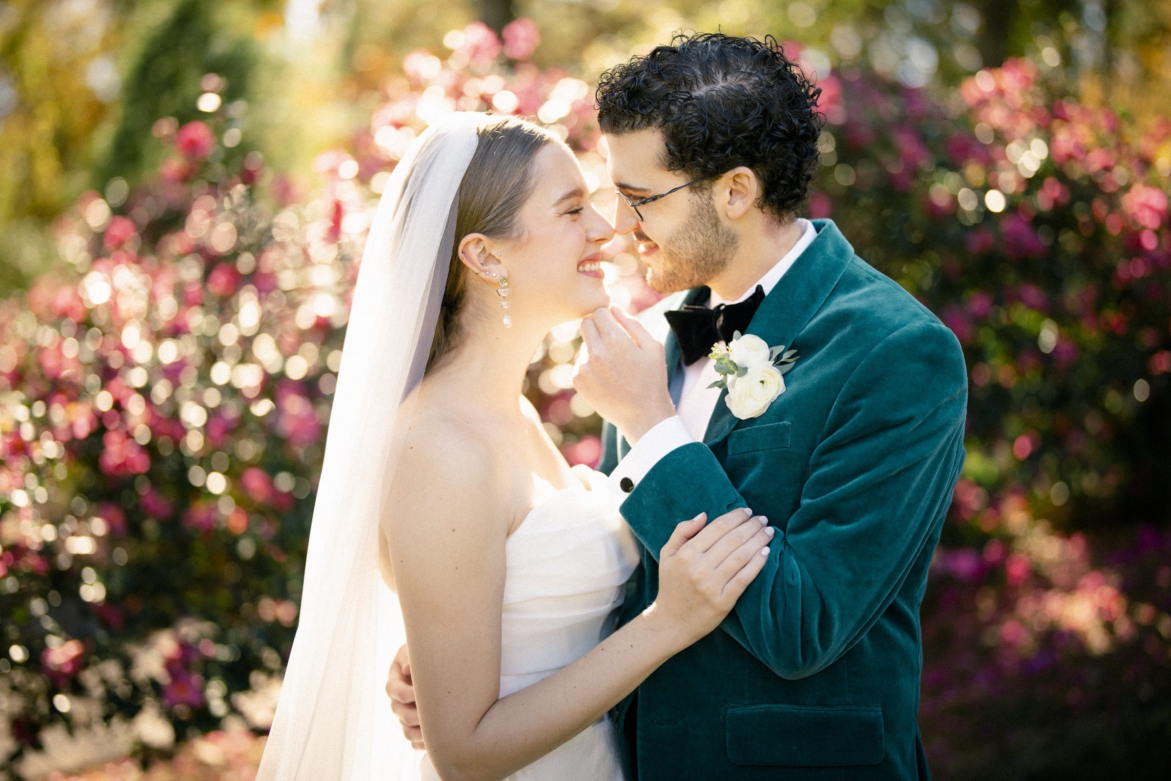 A bride and groom stand close, smiling at each other in an outdoor garden with colorful flowers. The bride wears a white dress and veil; the groom is in a green velvet suit jacket with a boutonniere.