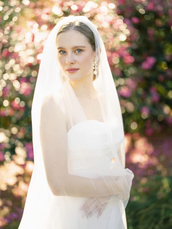 A bride in a white dress and veil stands outdoors, surrounded by vibrant pink flowers and greenery, with soft sunlight creating a dreamy, romantic atmosphere.