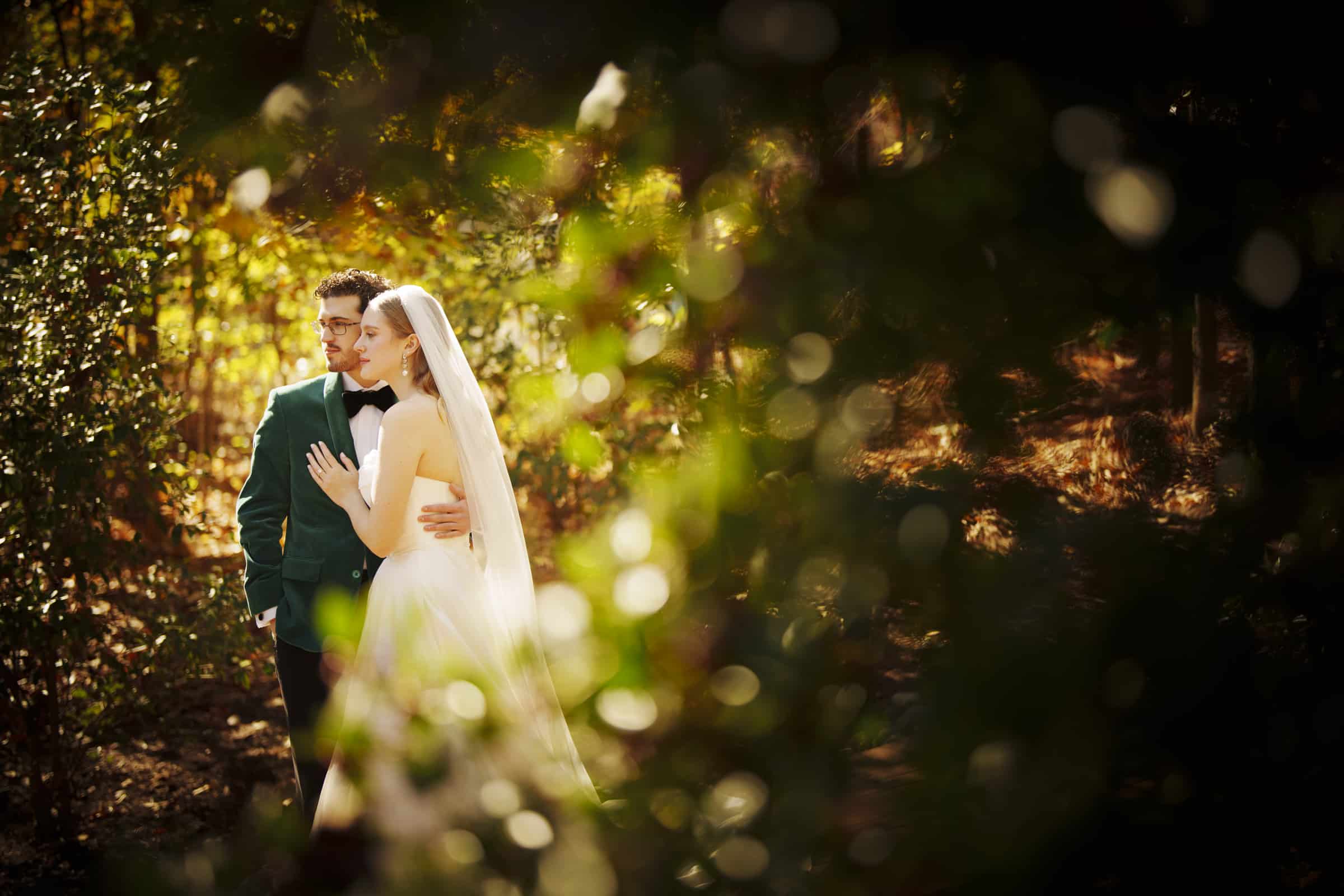 A bride in a white dress and veil embraces a groom in a green suit, standing closely together in a sunlit forest, surrounded by greenery and soft, blurred foliage in the foreground.