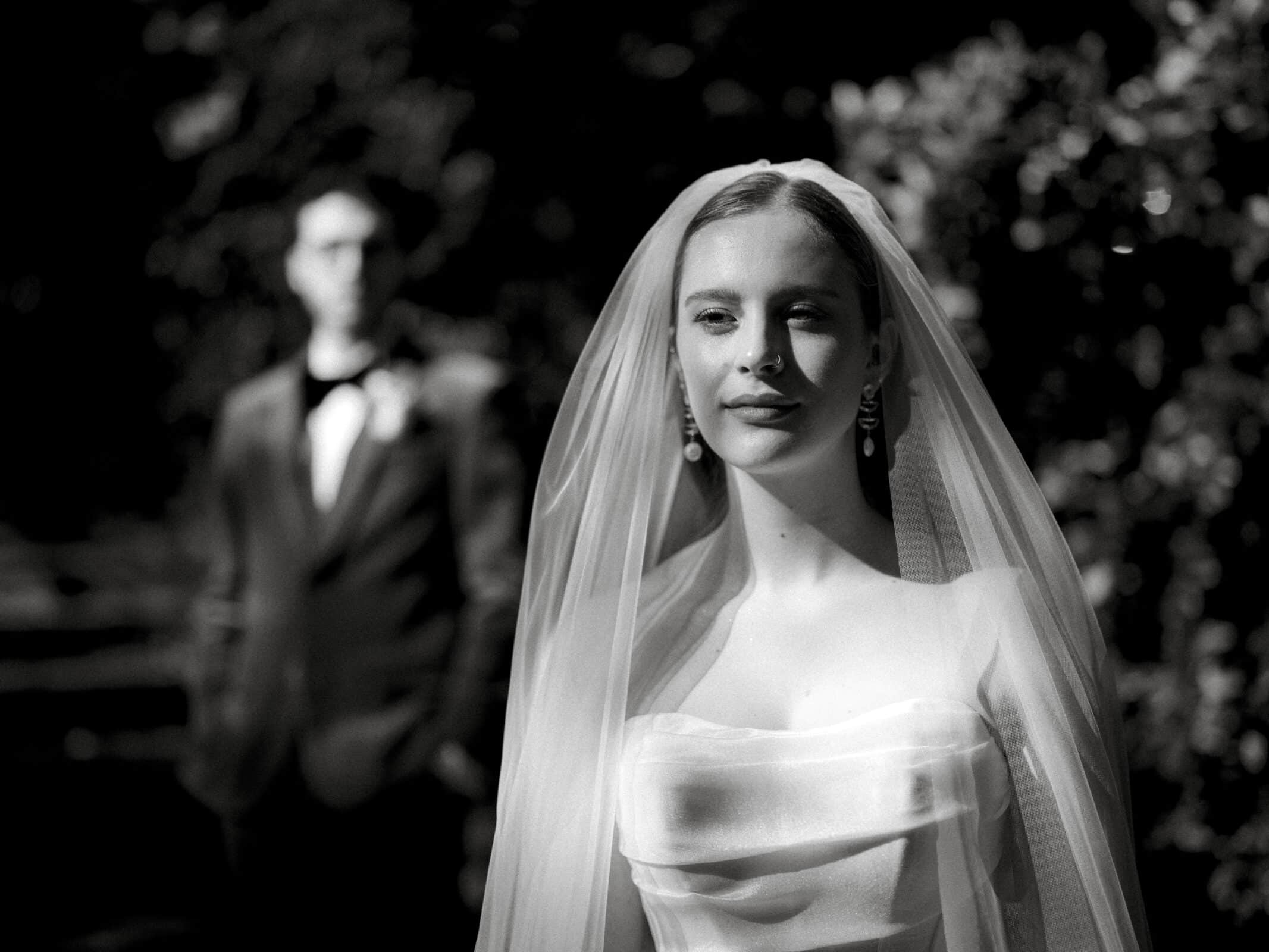 A black and white photo of a bride in a wedding dress and veil standing in the foreground, with a groom in a suit blurred in the background, outdoors.