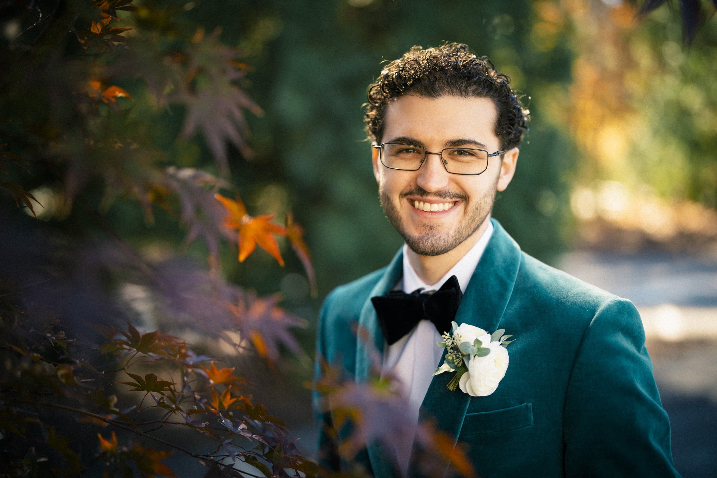 A man in a teal velvet suit with a black bow tie and white boutonniere smiles outdoors, framed by autumn leaves, with soft natural light and a blurred green background.
