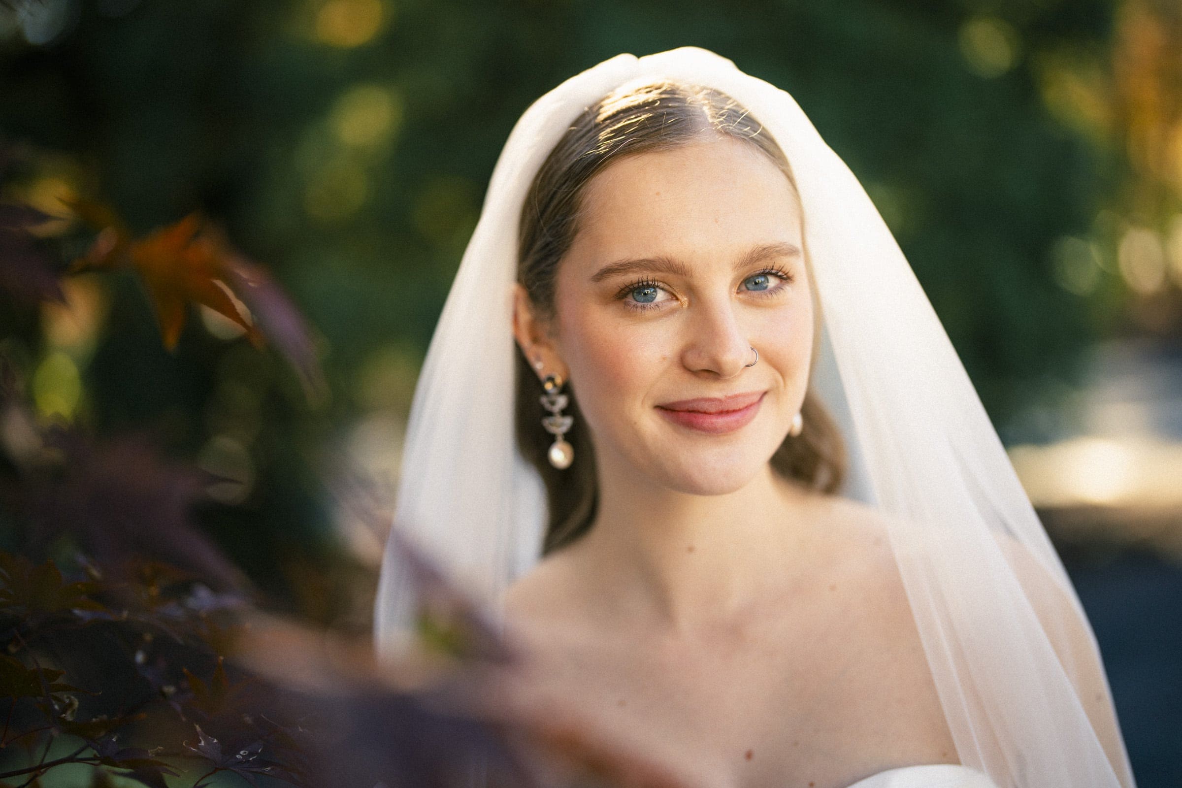 A bride with blue eyes and light brown hair smiles softly. She wears a white veil, drop earrings, and a strapless dress, standing outdoors with sunlight and greenery in the background.