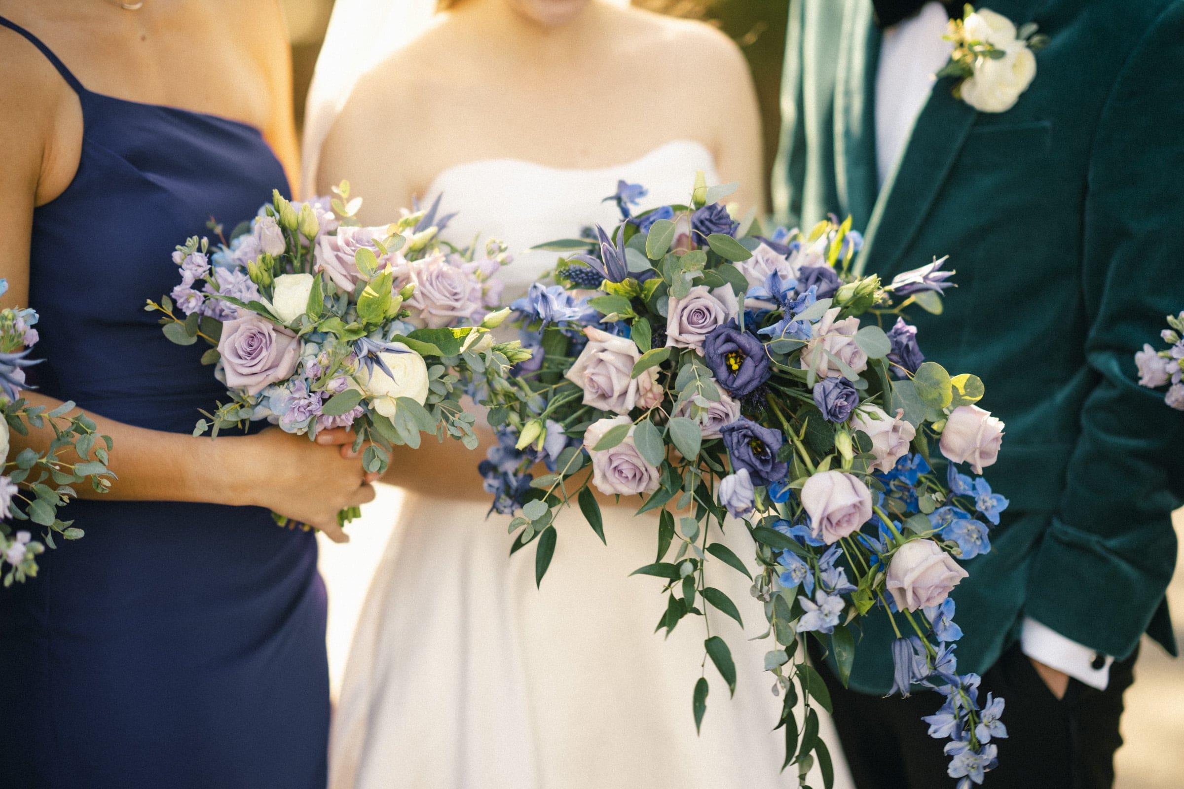 Three people stand close together holding bouquets of purple and blue flowers; the central figure wears a white dress, another wears a dark blue dress, and the third wears a green suit with a boutonniere.