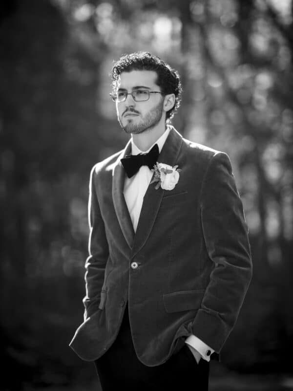 A man wearing glasses, a bow tie, and a suit jacket with a boutonniere stands outdoors with his hands in his pockets, looking thoughtful. The background is blurred with sunlight filtering through trees.