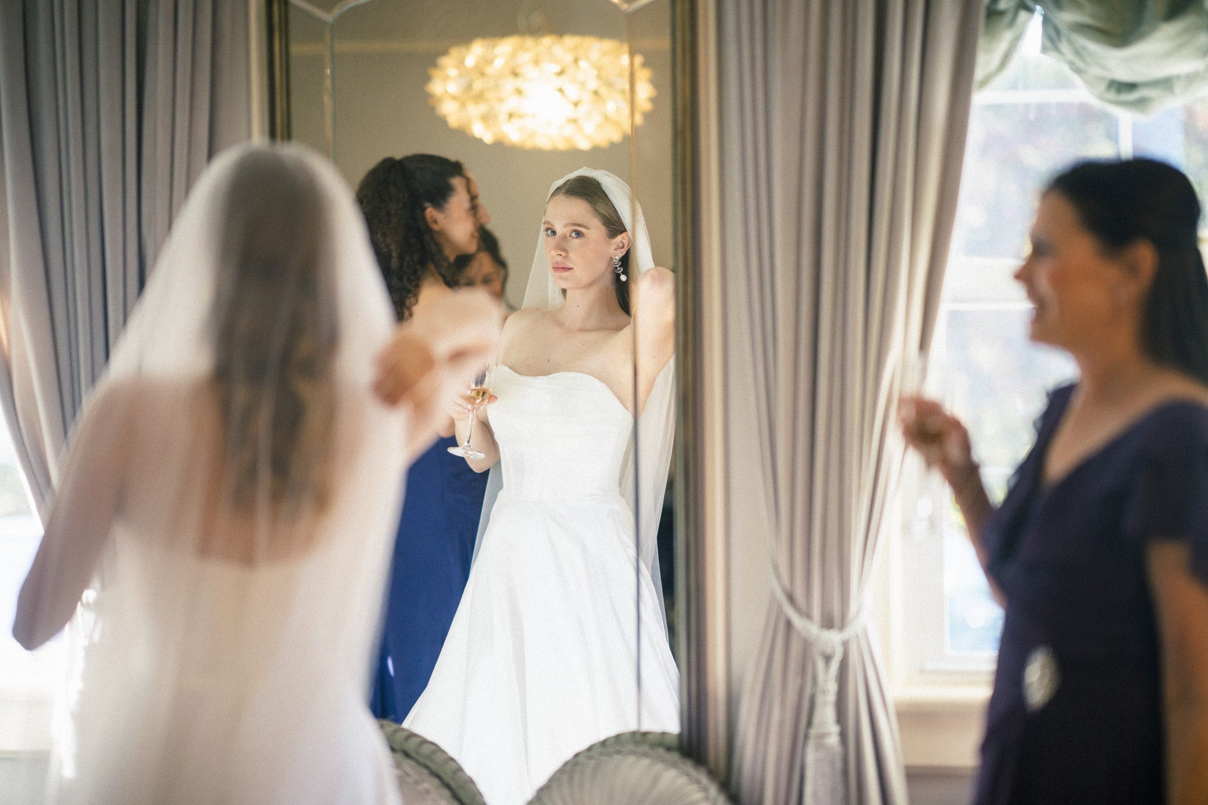 A bride in a white wedding dress looks at her reflection in a large mirror, holding a glass. Two women, one blurred in the foreground and one beside her, are also present in the elegant room with light curtains.