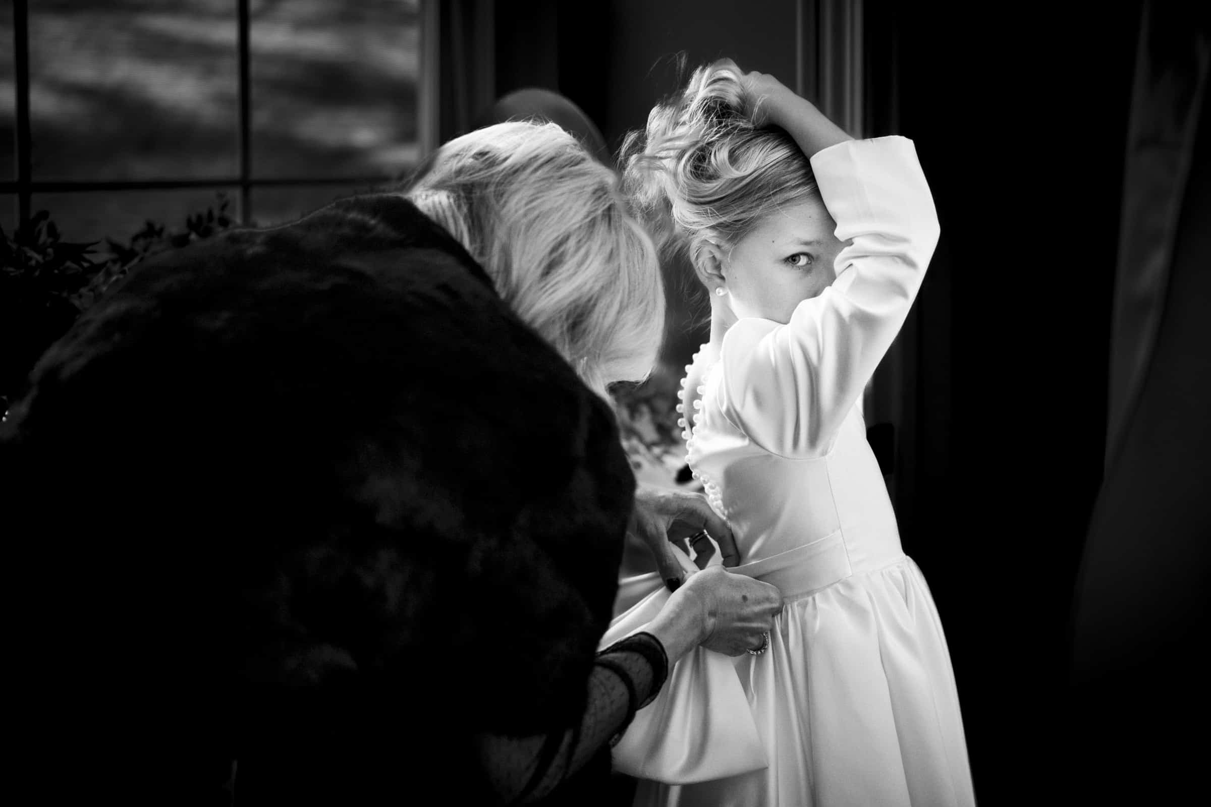 A young girl in a white dress adjusts her hair while an older woman fastens the back of her dress. The scene is indoors with soft lighting, and the girl looks directly at the camera.