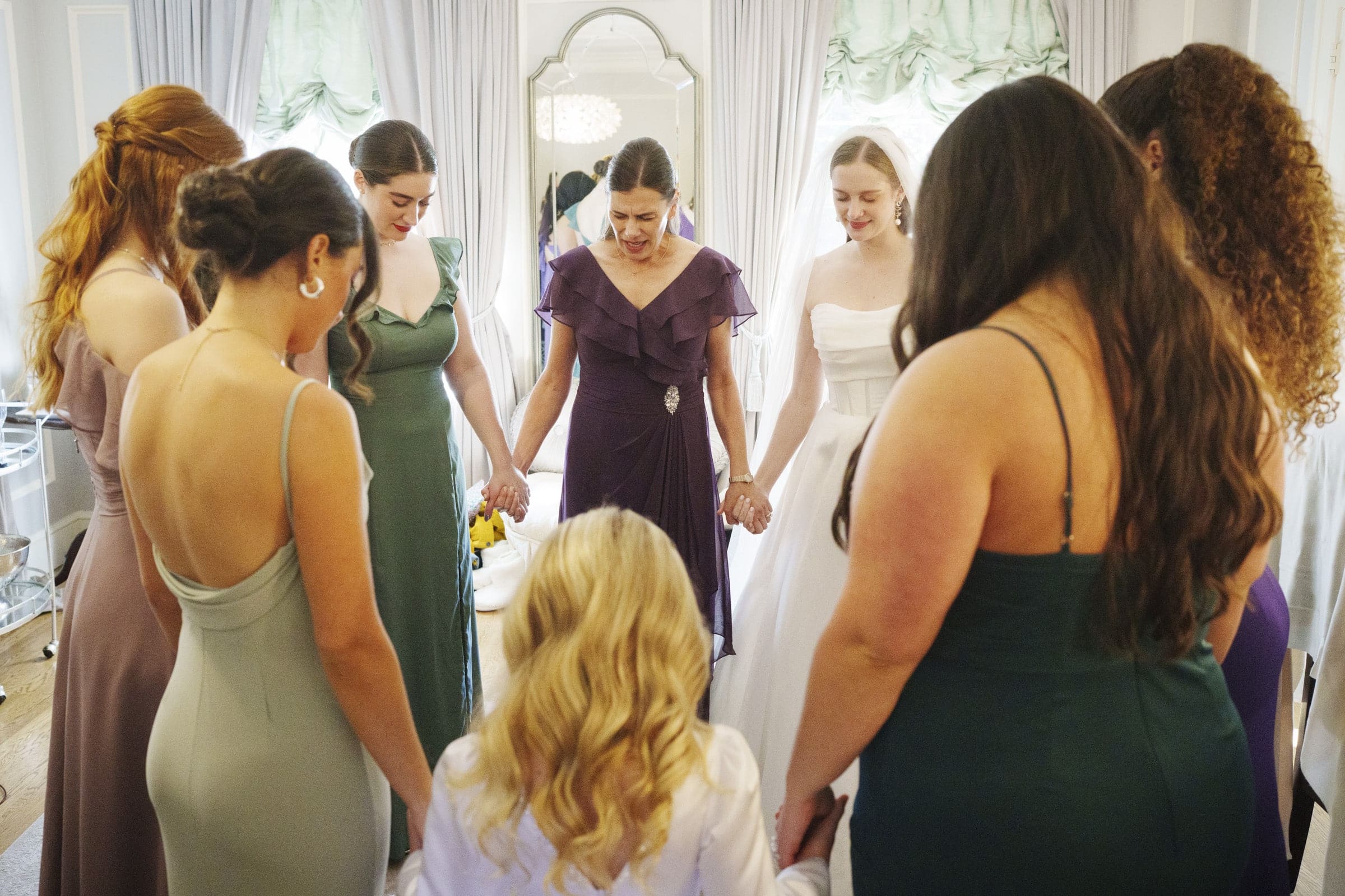 A group of women in formal dresses stand in a circle, holding hands and bowing their heads, appearing to pray or share a moment of reflection in a softly lit room.