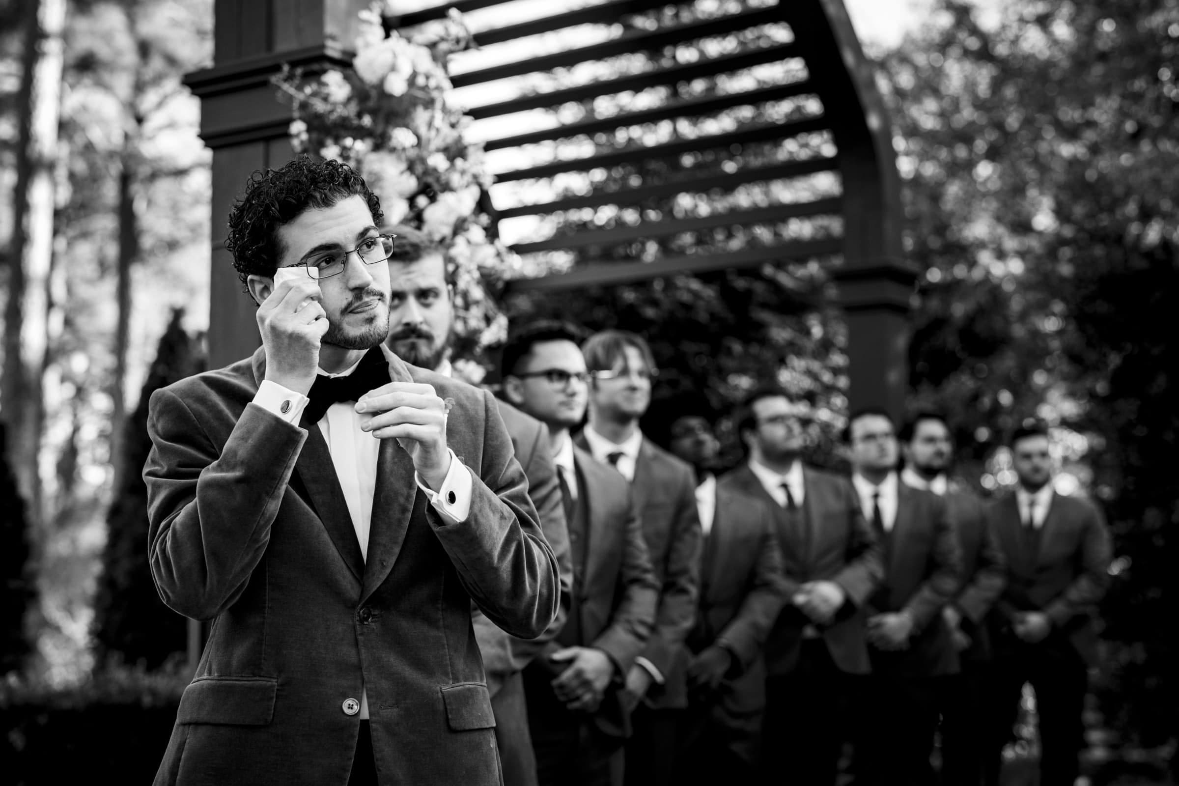 A groom in a suit wipes his eyes with a tissue during an outdoor wedding ceremony, while several groomsmen stand in a line behind him under a wooden archway decorated with flowers.