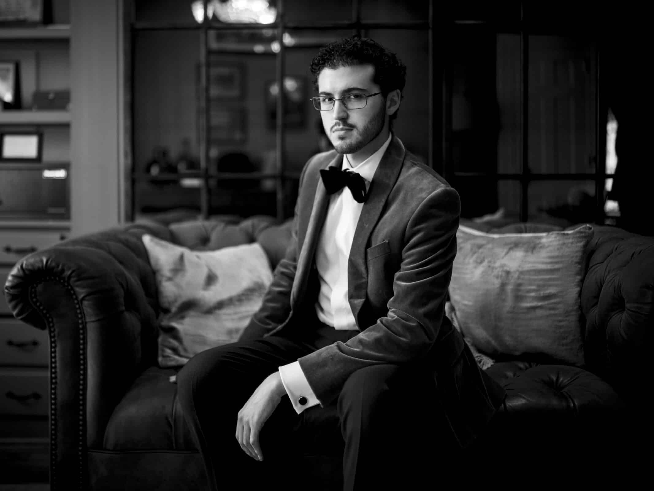 A man in a tuxedo and bow tie sits on a tufted sofa in a dimly lit room, looking thoughtfully to the side. The image is in black and white, creating a classic, elegant atmosphere.