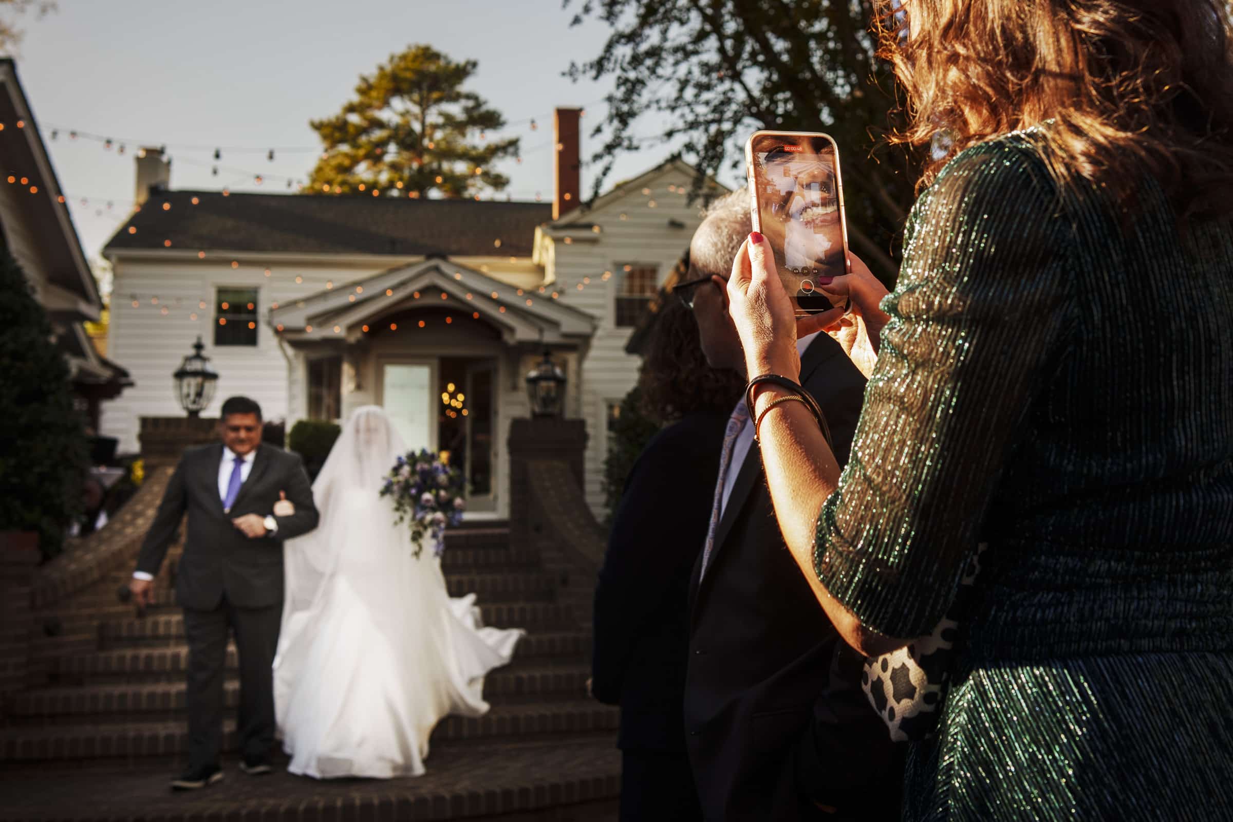 A woman in a green dress holds up her phone to take a photo or video as a bride in a white gown and a man in a suit walk down steps outside a house decorated with string lights.