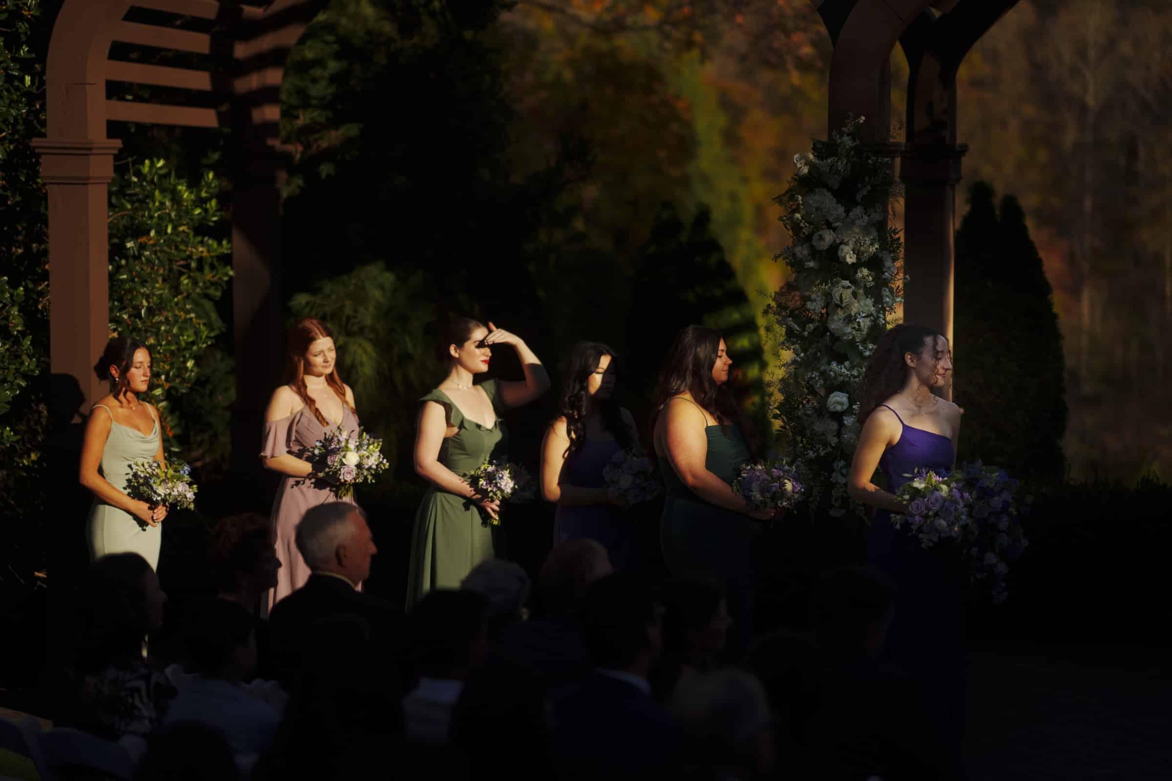 Six bridesmaids in colorful dresses stand in a row holding bouquets, sunlight casting dramatic shadows. They face right, with one shielding her eyes, surrounded by greenery and floral decorations at an outdoor ceremony.