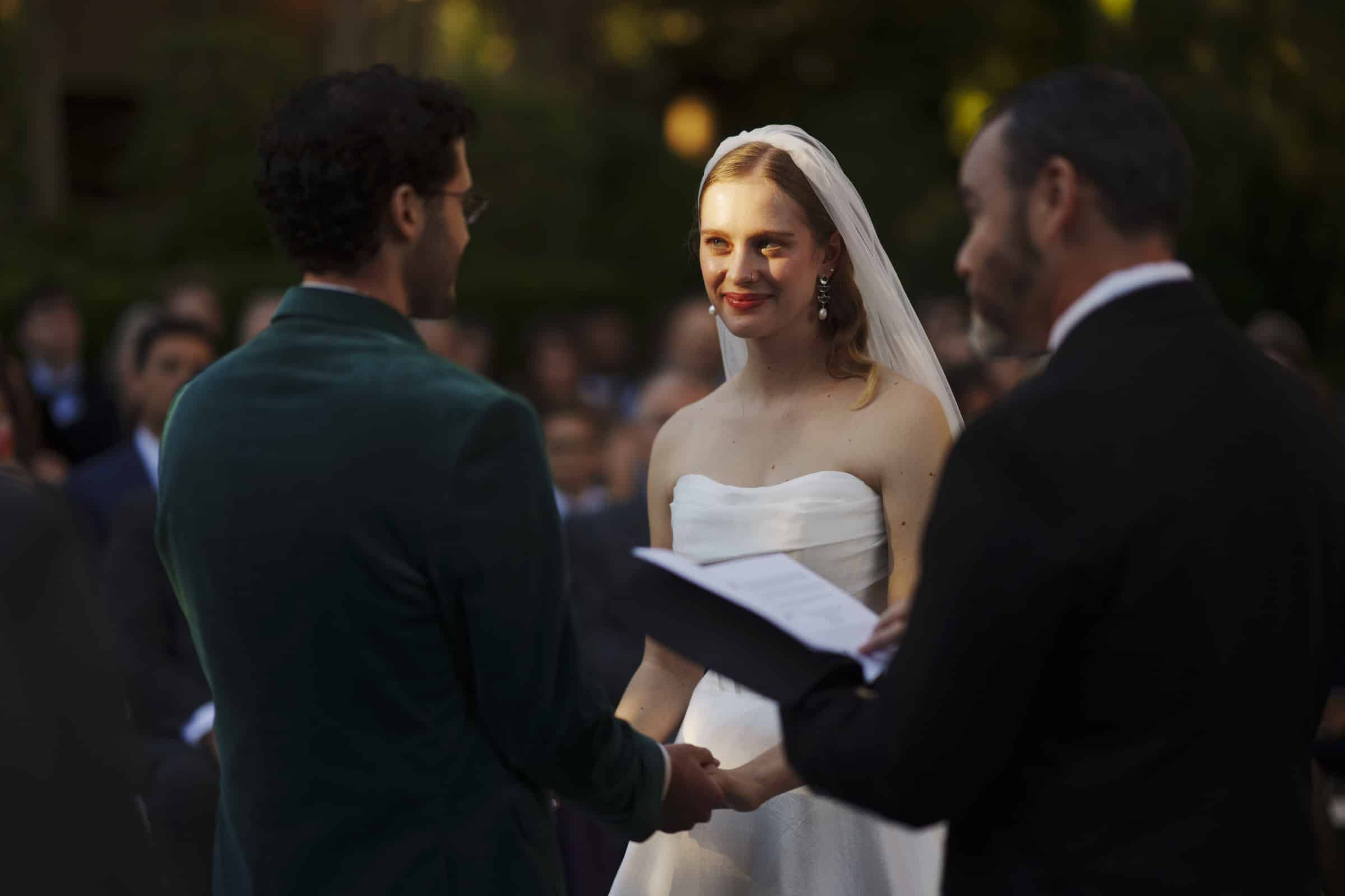 A bride in a white dress and veil smiles at her partner during an outdoor wedding ceremony, holding hands, while an officiant reads from a book.