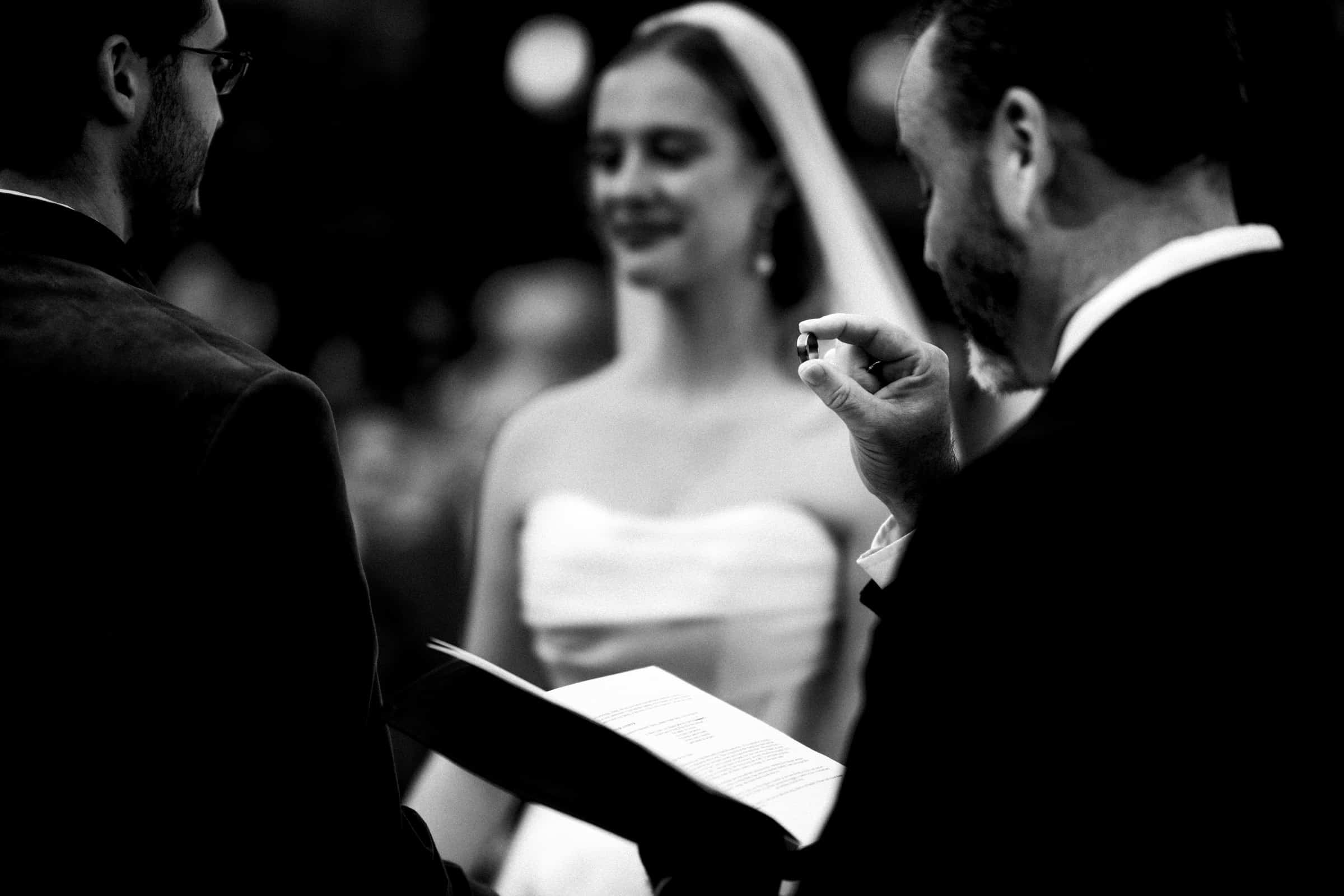 A black and white photo of a wedding ceremony shows a bride in a strapless dress smiling, while an officiant holds a ring and a book, with another person standing nearby.