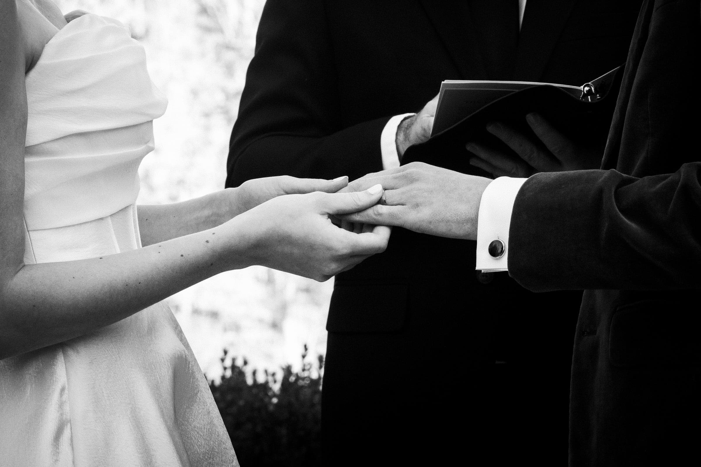A bride and groom exchange rings during a wedding ceremony, with an officiant holding a book in the background. The image is in black and white and focuses on their hands.