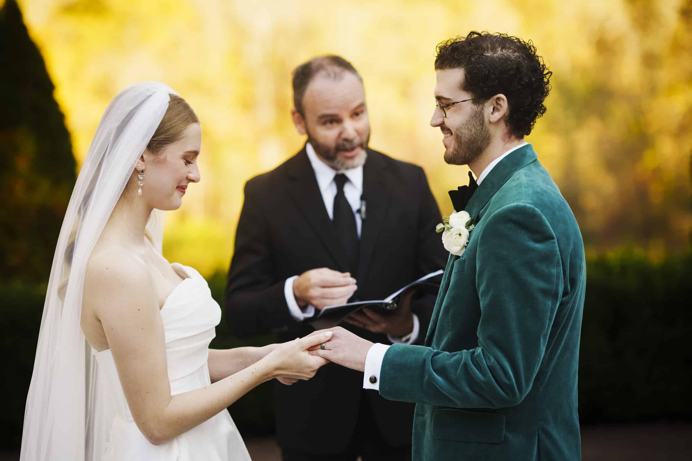 A bride and groom smile at each other while holding hands during an outdoor wedding ceremony, as an officiant stands behind them speaking and holding a book.