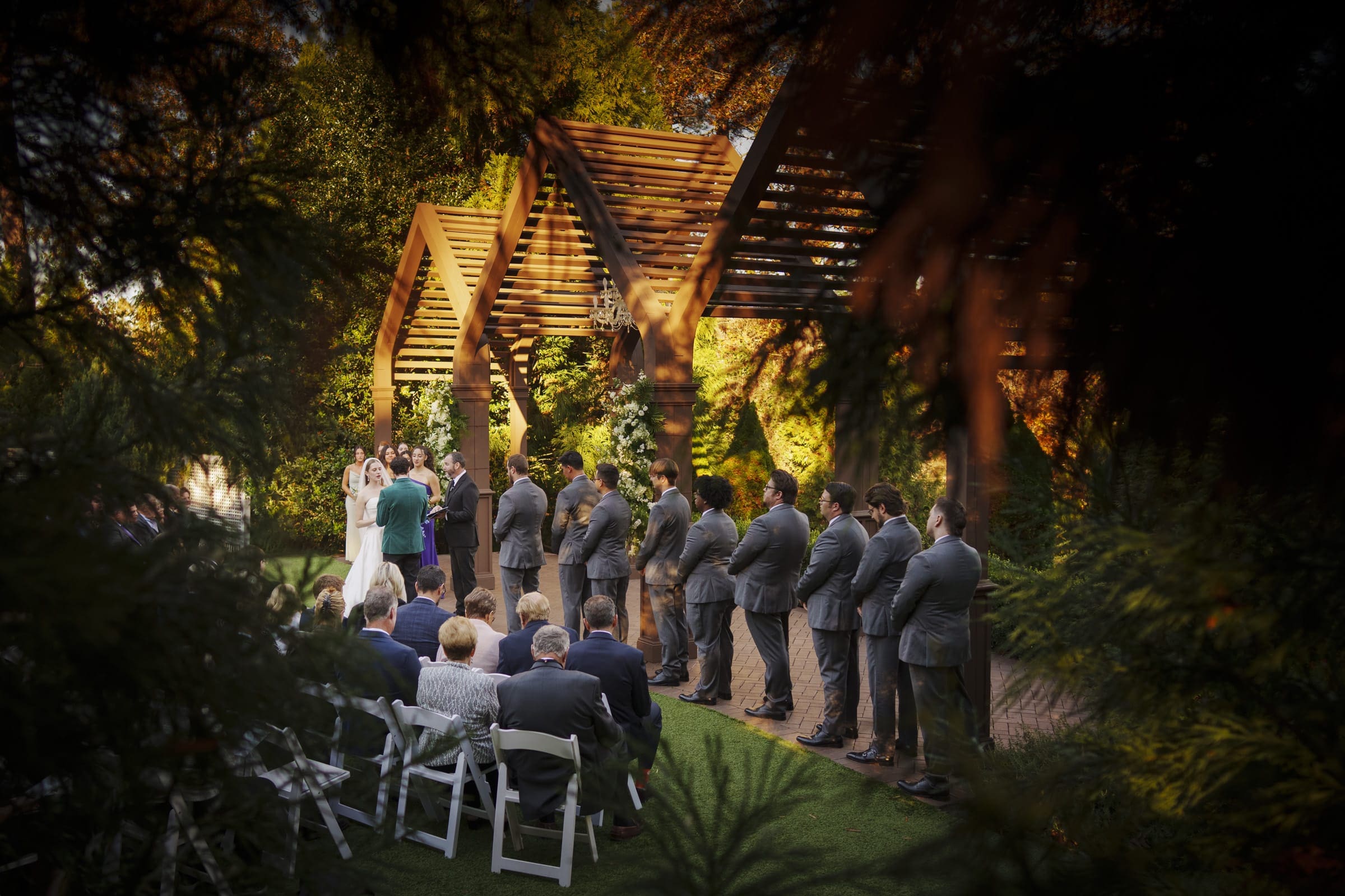 A wedding ceremony takes place outdoors under wooden arches, with the bride and groom at the altar, groomsmen in suits lined up, and guests seated on white chairs, surrounded by greenery and warm sunlight.