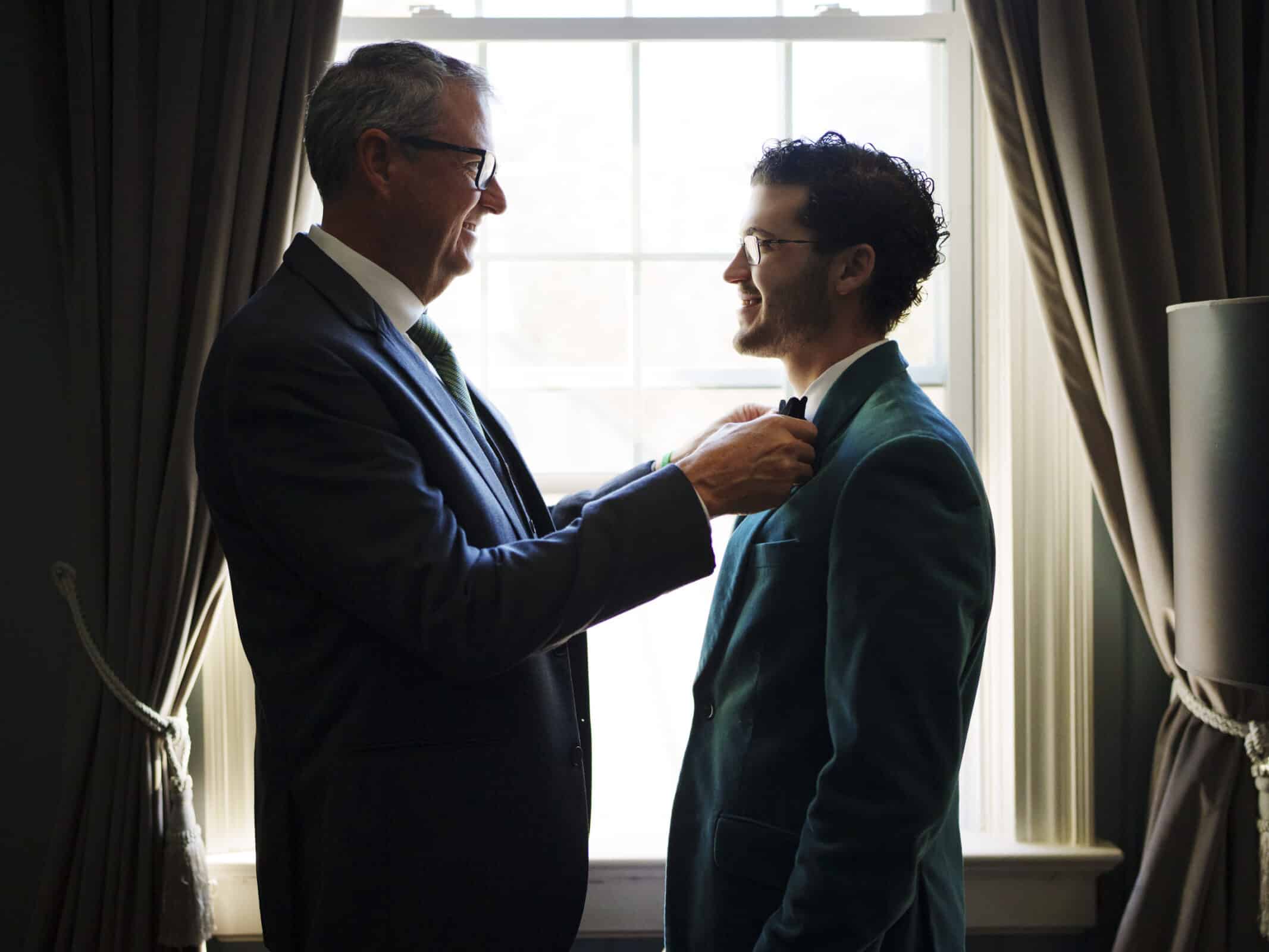 Two men in suits stand by a window with curtains. The older man is adjusting the younger mans bow tie, both smiling warmly at each other. Soft natural light fills the room.