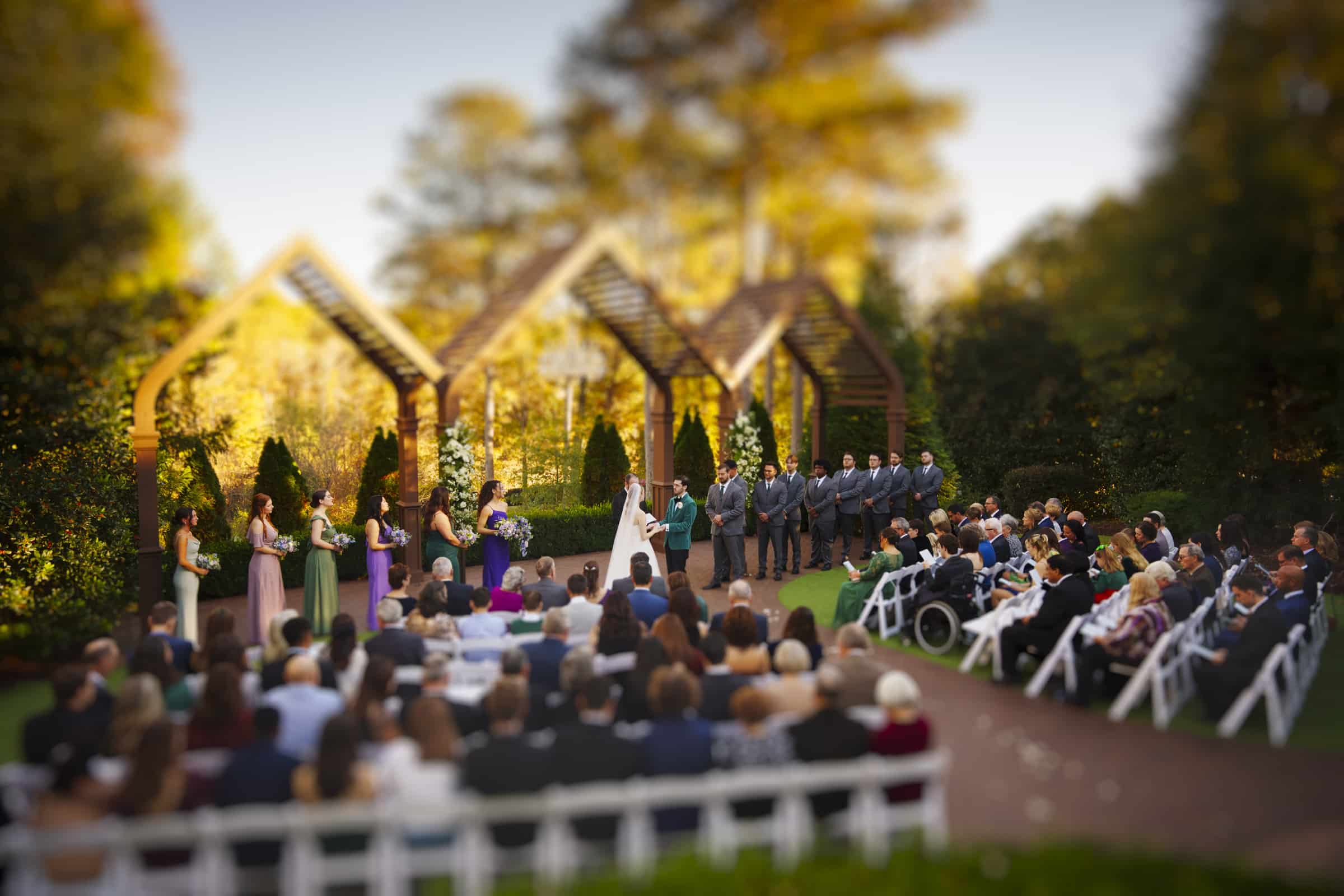 A bride and groom stand at an outdoor wedding altar surrounded by their wedding party, while guests seated in white chairs watch the ceremony in a garden setting.