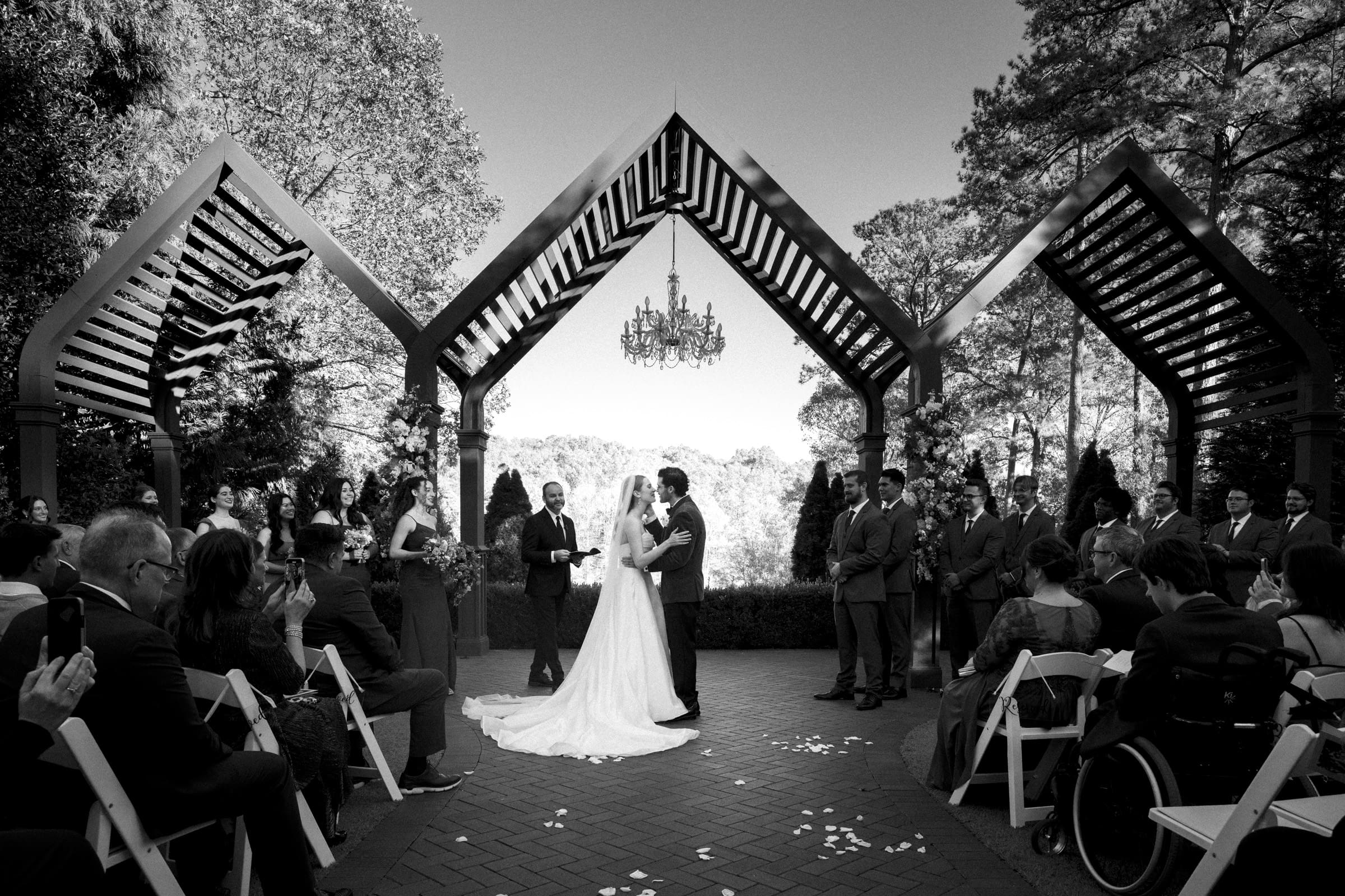 A bride and groom stand under a decorative arch, holding hands during their outdoor wedding ceremony, surrounded by guests seated on white chairs and their wedding party. The scene is in black and white.