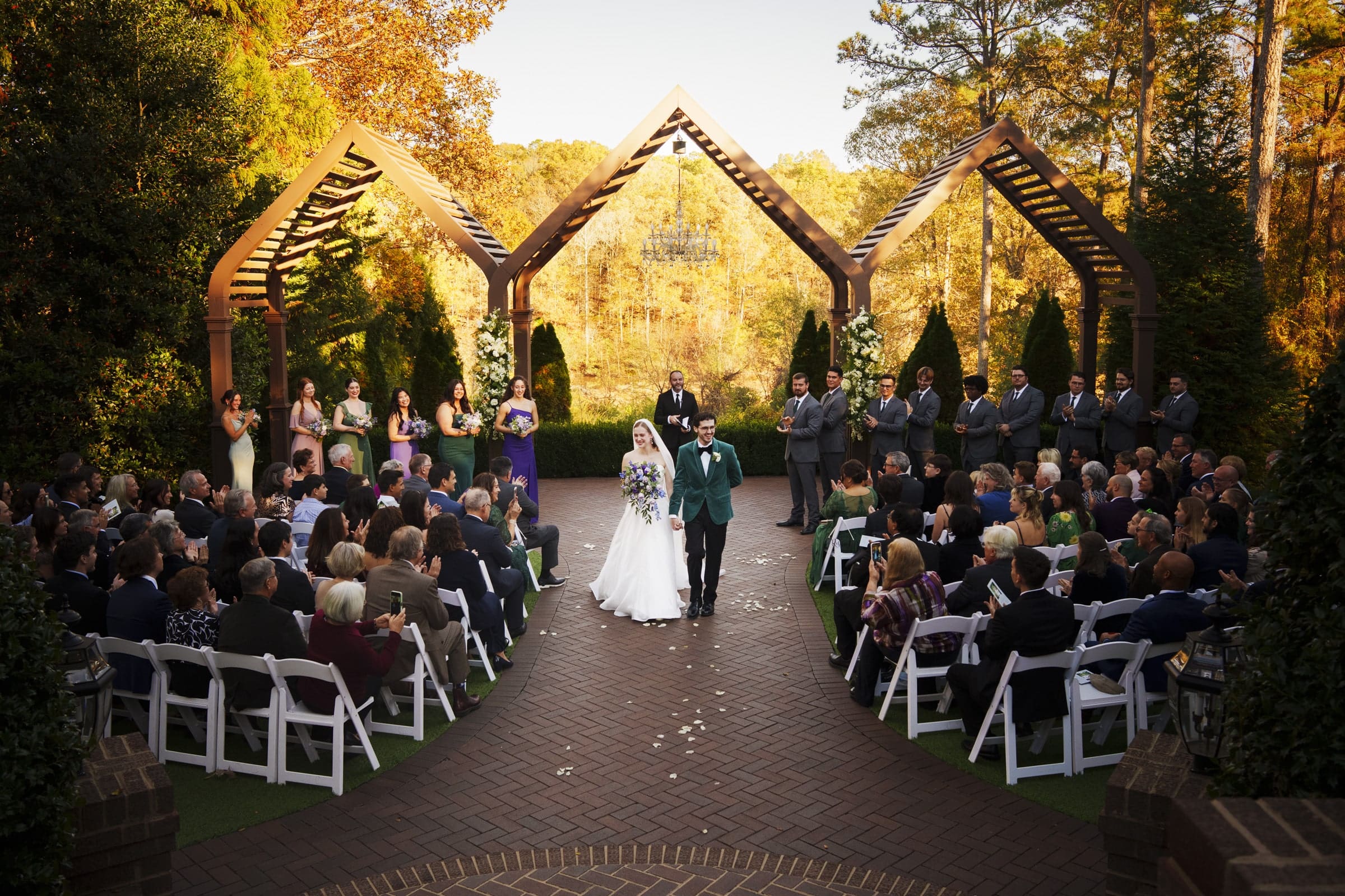 A bride and groom walk down the aisle at an outdoor wedding ceremony, surrounded by seated guests and a wedding party standing under wooden arches, with trees and golden sunlight in the background.