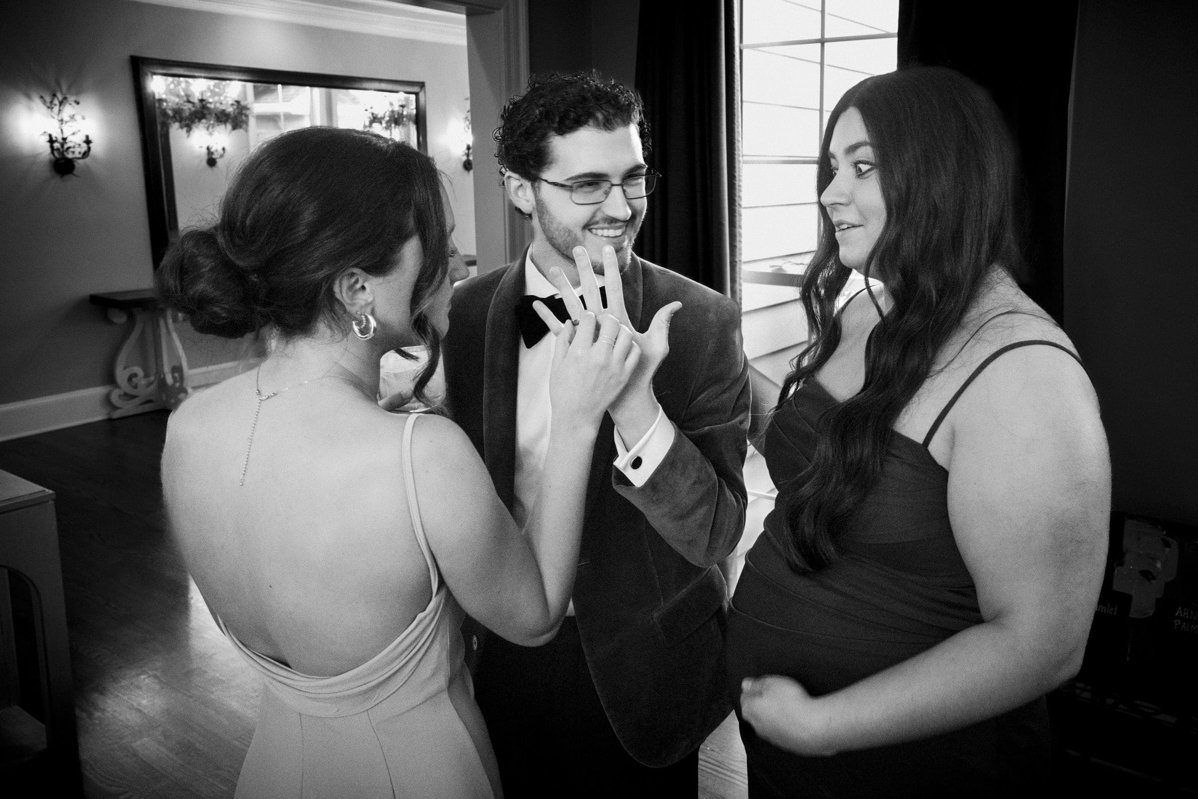 Three people stand indoors, with one woman showing her hand and ring to a man and another woman. They are smiling and appear to be celebrating an engagement. The setting is elegant, with wall sconces and a large mirror.
