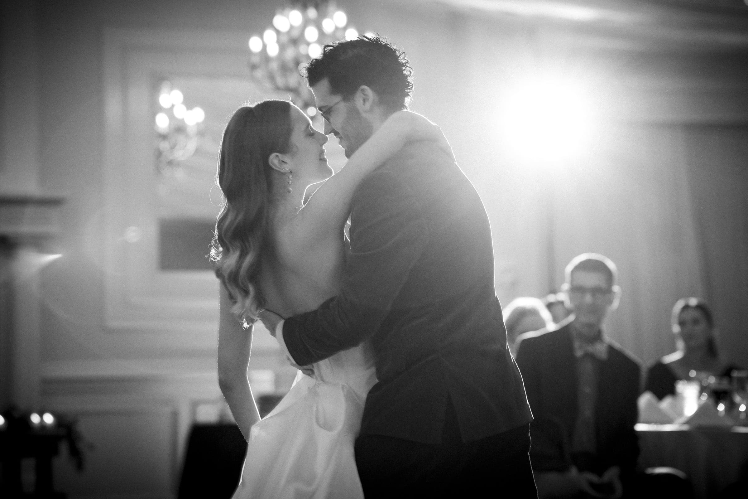 A bride and groom share a joyful first dance in a softly lit ballroom, gazing into each other’s eyes and smiling. Guests watch from tables in the background. The scene is elegant and romantic.