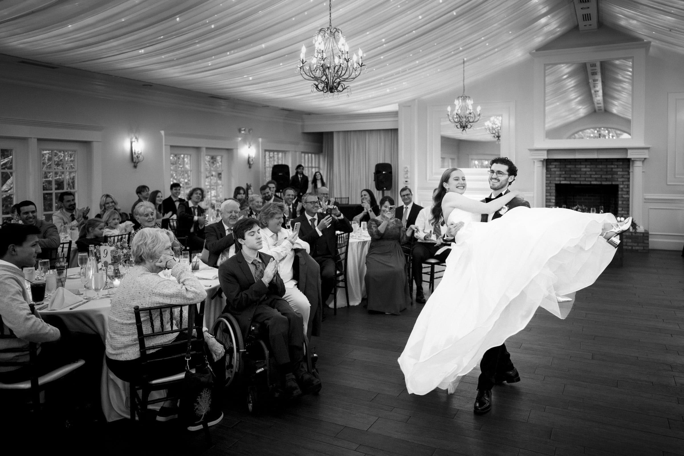 A groom lifts and twirls his bride on the dance floor as wedding guests seated at tables watch and applaud in a decorated, elegant reception hall.