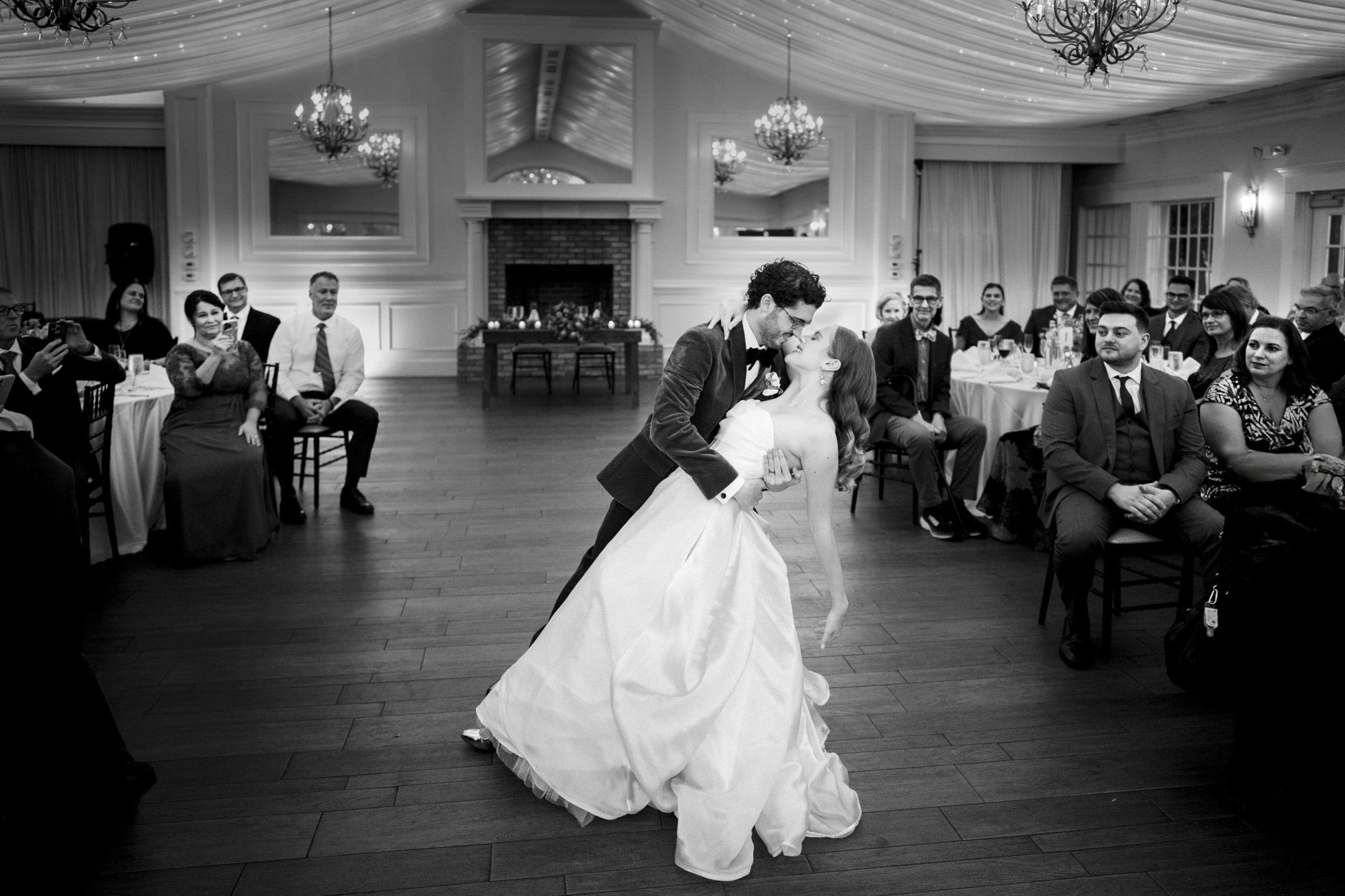A bride and groom share a romantic dip during their first dance at a wedding reception, while guests seated at tables smile and watch in a beautifully decorated hall.
