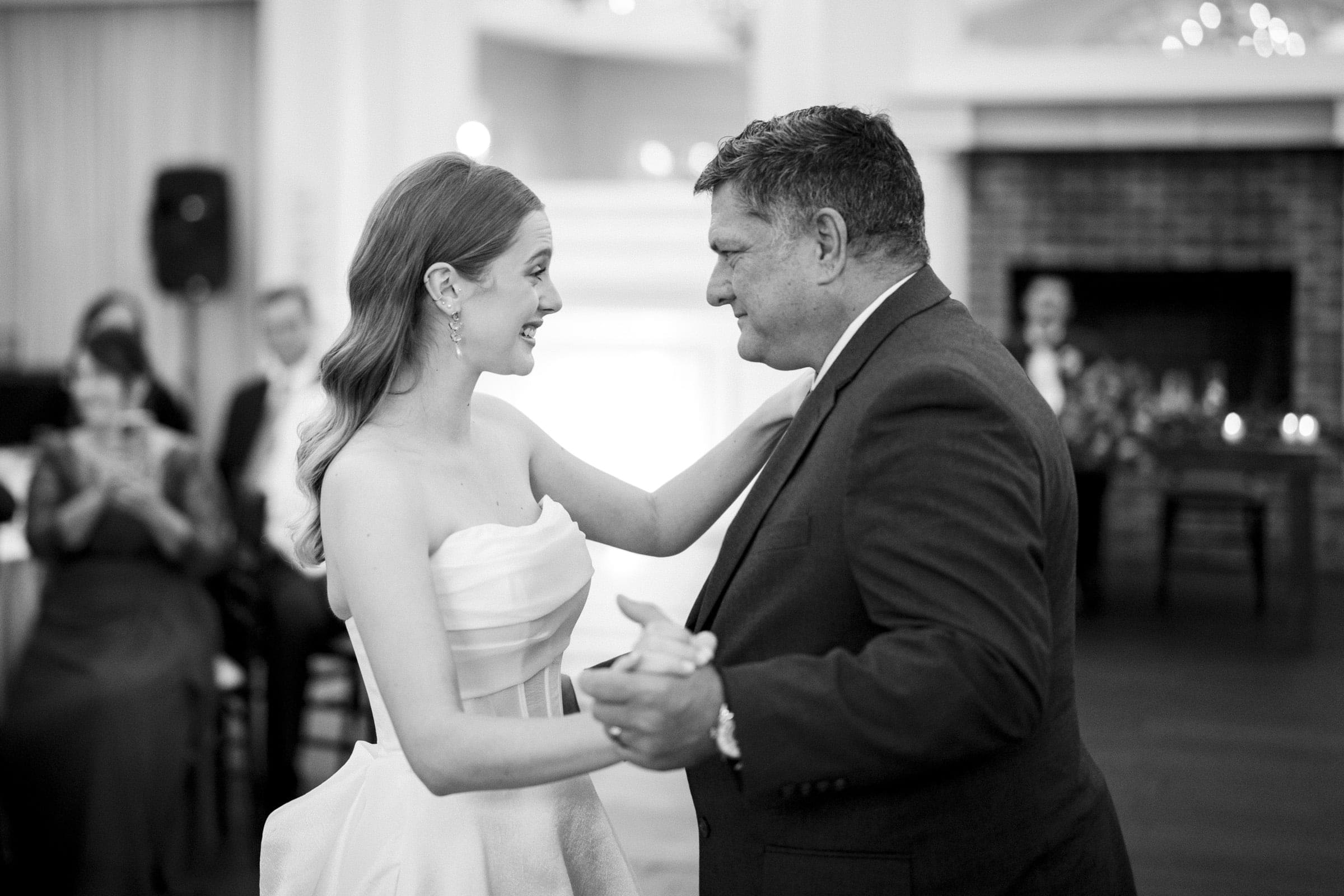 A bride in a white dress smiles as she dances with an older man in a suit at a wedding reception. Guests seated in the background watch the pair. The image is in black and white.