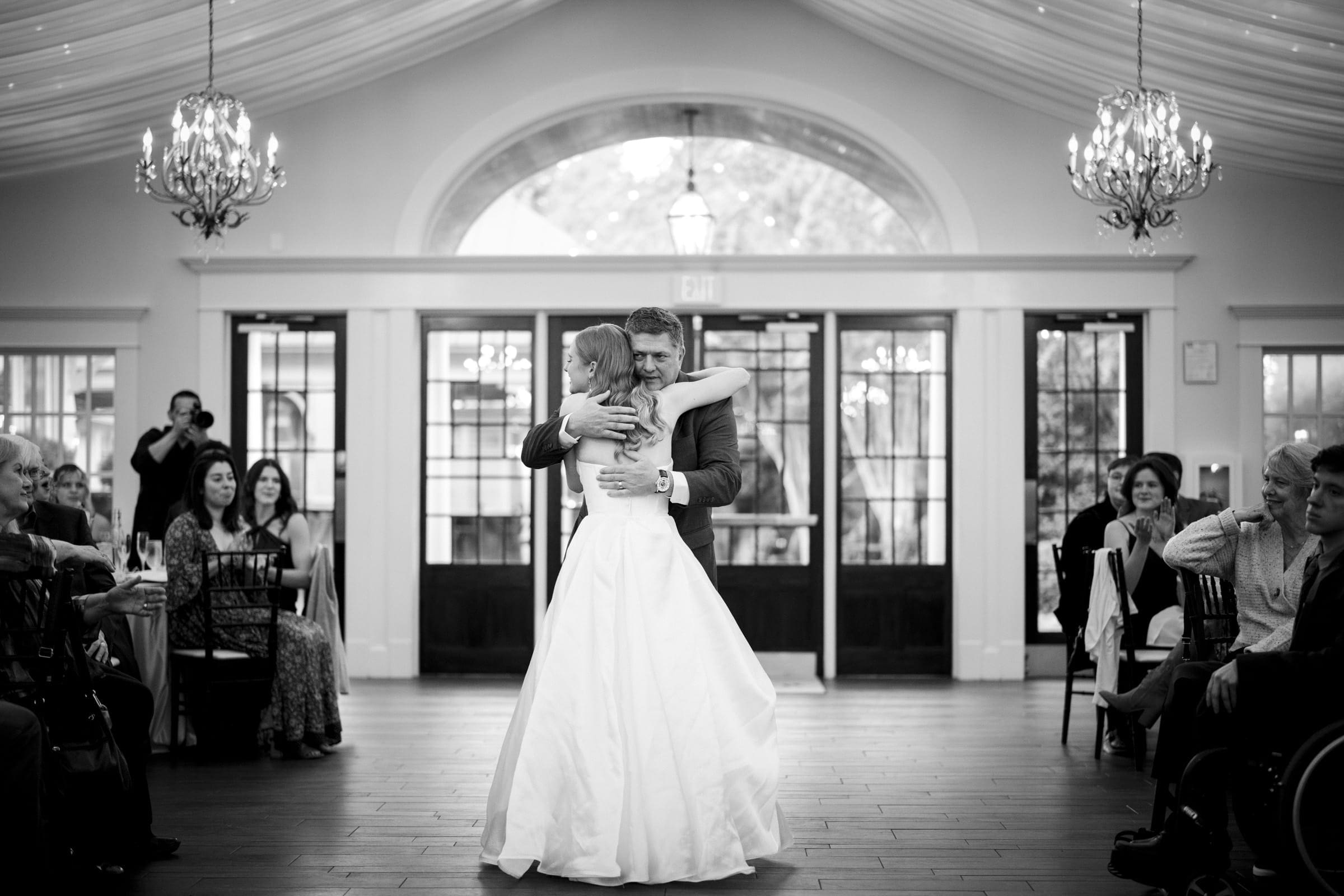 A bride in a white gown and an older man in a suit share a heartfelt embrace on a dance floor at a wedding reception while guests watch from their tables in an elegant, chandelier-lit venue.