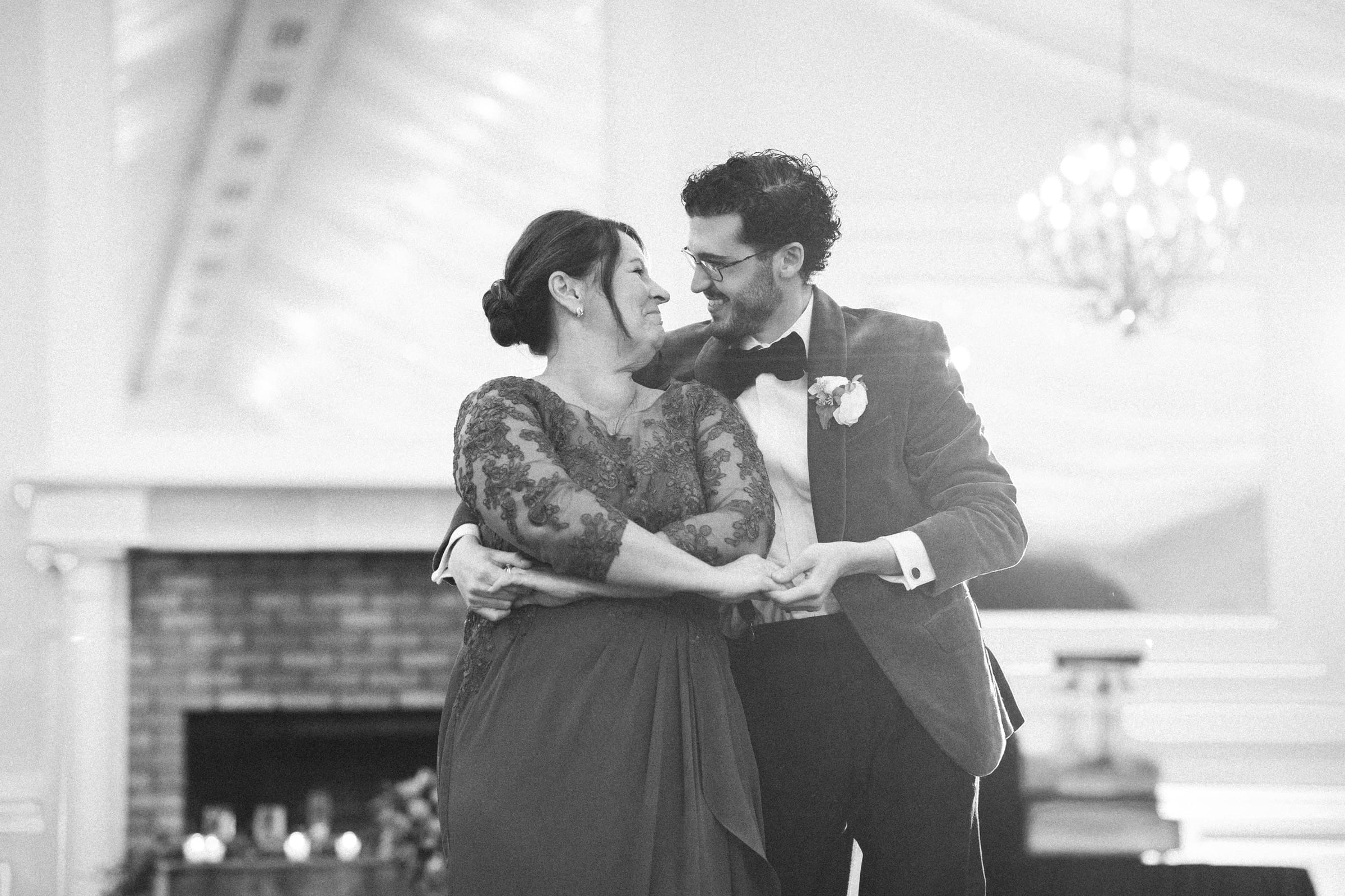 A couple dressed formally, smiling and dancing together in an elegant, softly lit room with a chandelier and fireplace in the background.