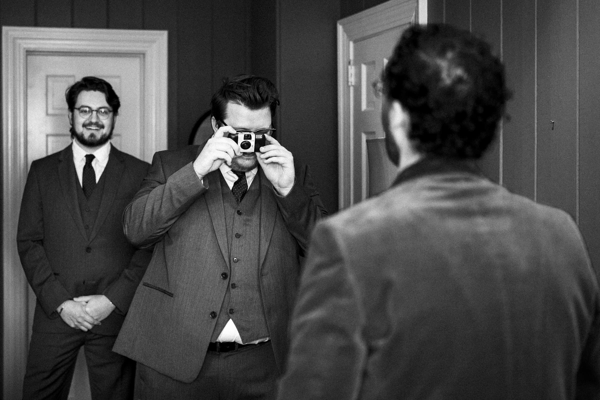 Two men in suits stand indoors; one stands in front of a door, smiling, while the other takes a photo with a camera, his reflection visible in a mirror. The scene is in black and white.