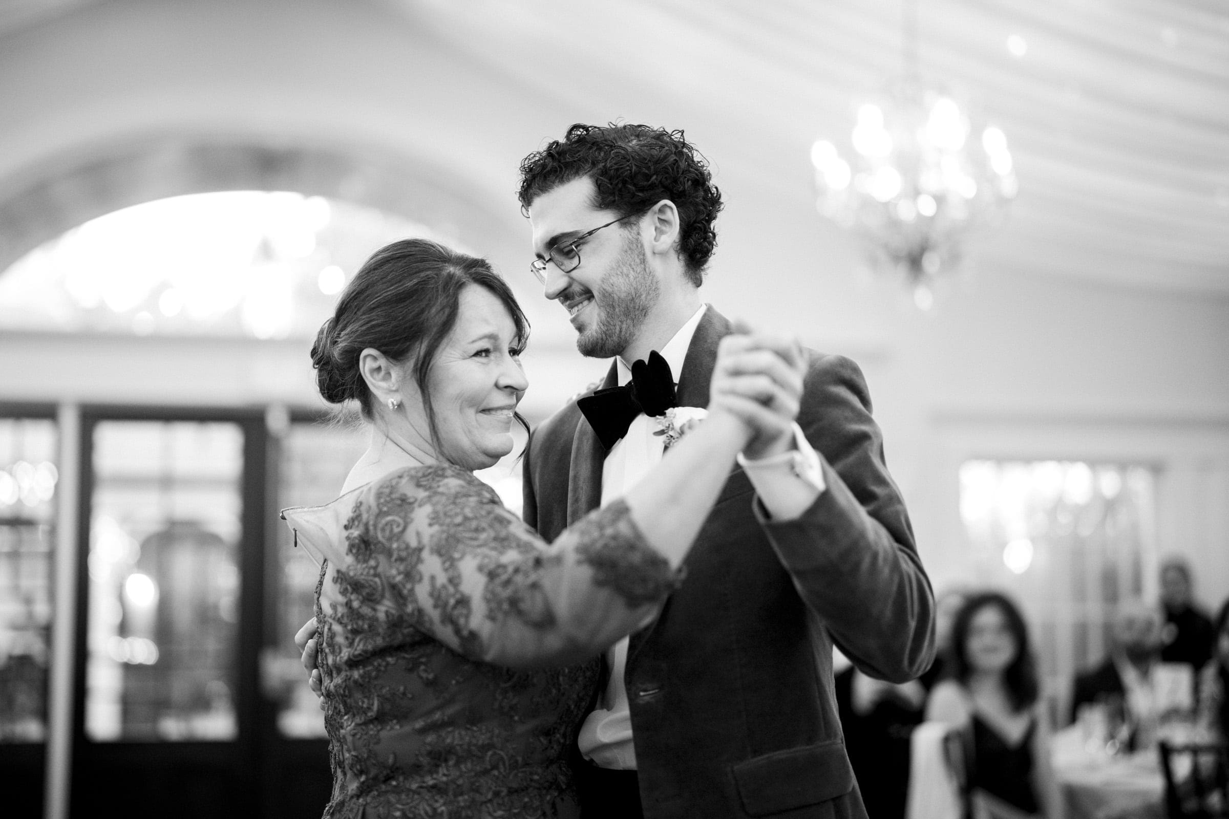 A smiling man in a suit and bow tie dances with a woman in a lace dress at a formal event, likely a wedding. They look at each other joyfully under a chandelier, with guests seated in the background.