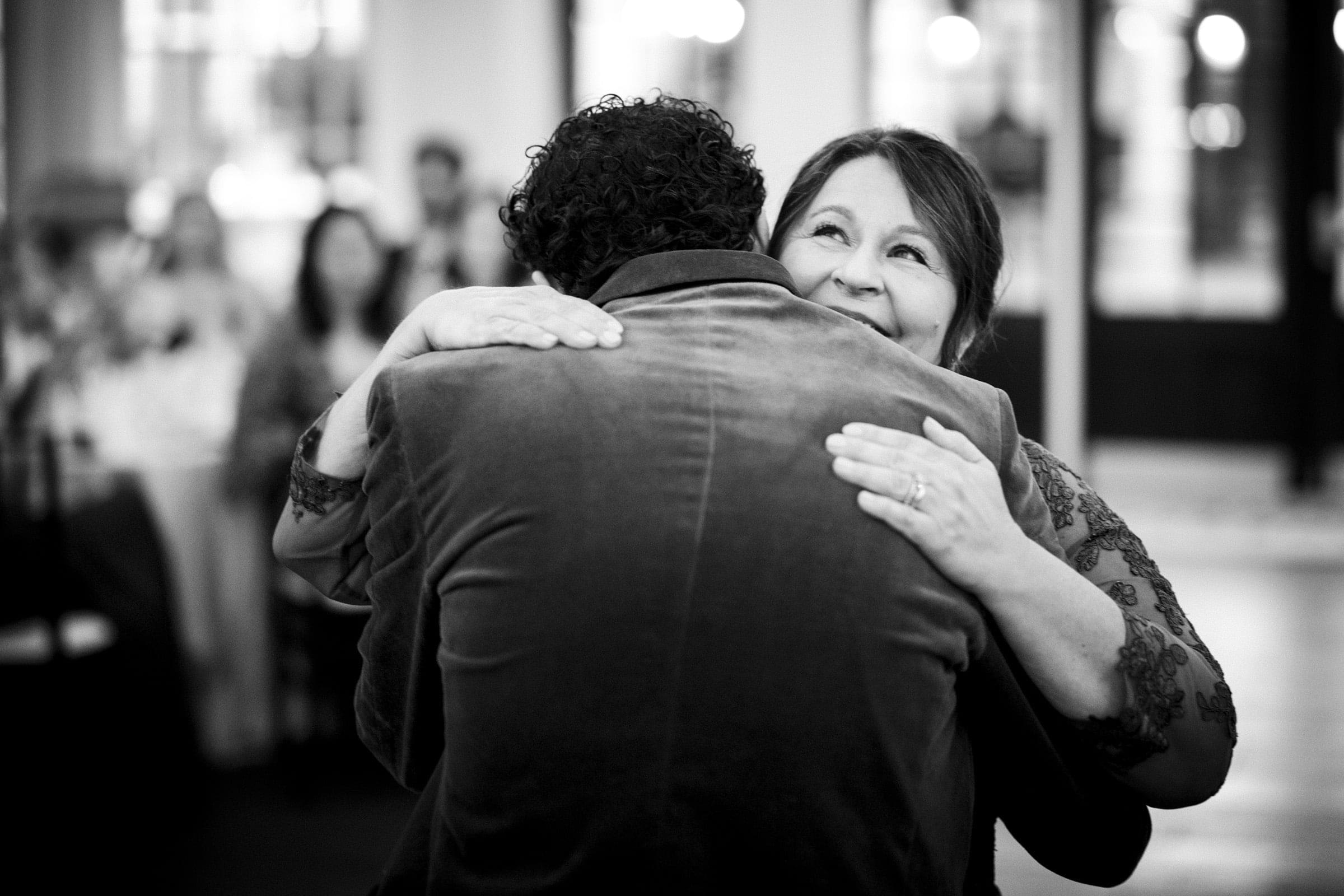 A woman smiles warmly while hugging a man in a velvet jacket at what appears to be a celebratory event, with blurred guests in the background. The image is in black and white.