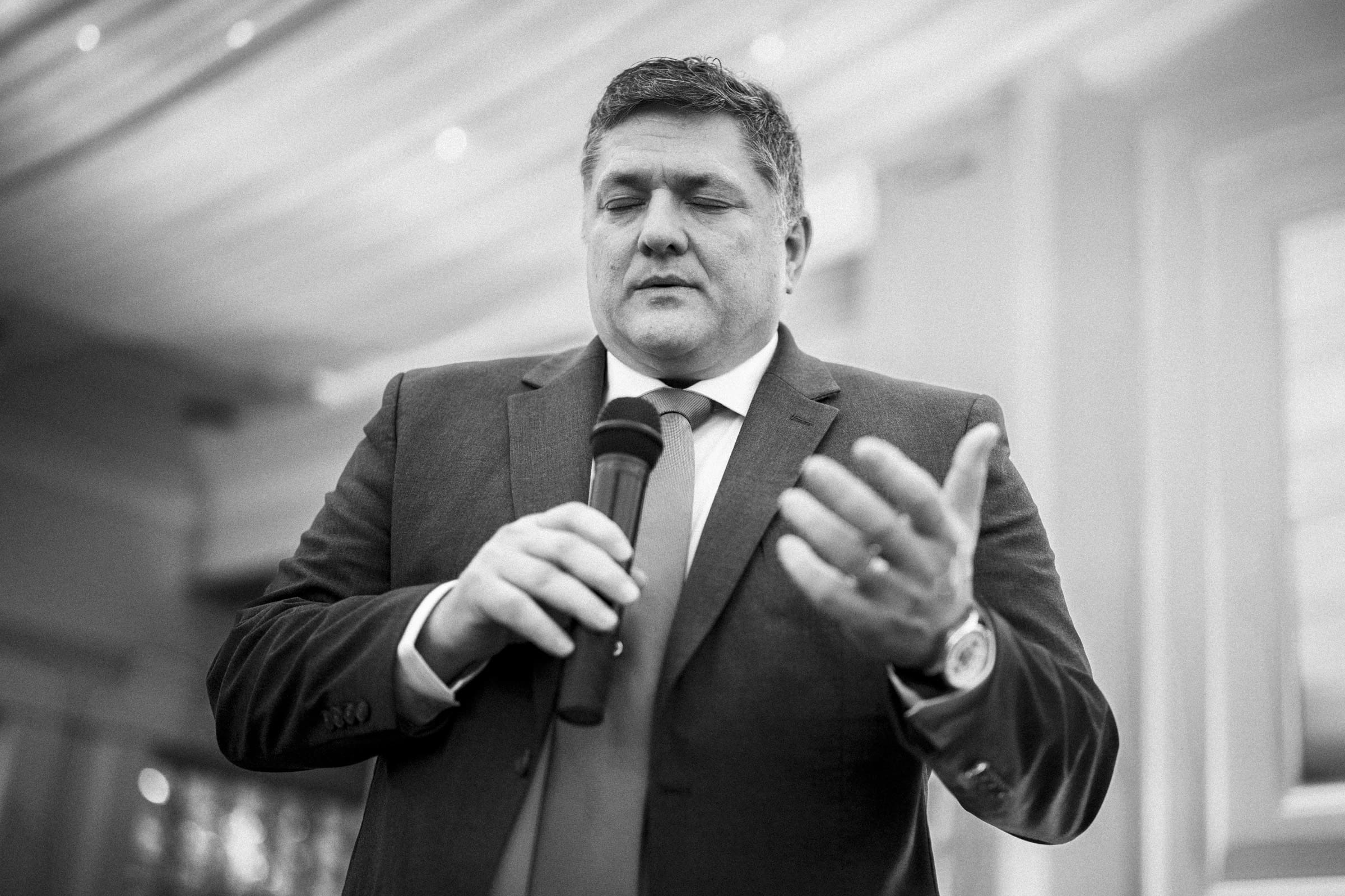 A man in a suit speaking into a microphone stands indoors, gesturing with his hand, with his eyes closed. The image is in black and white.